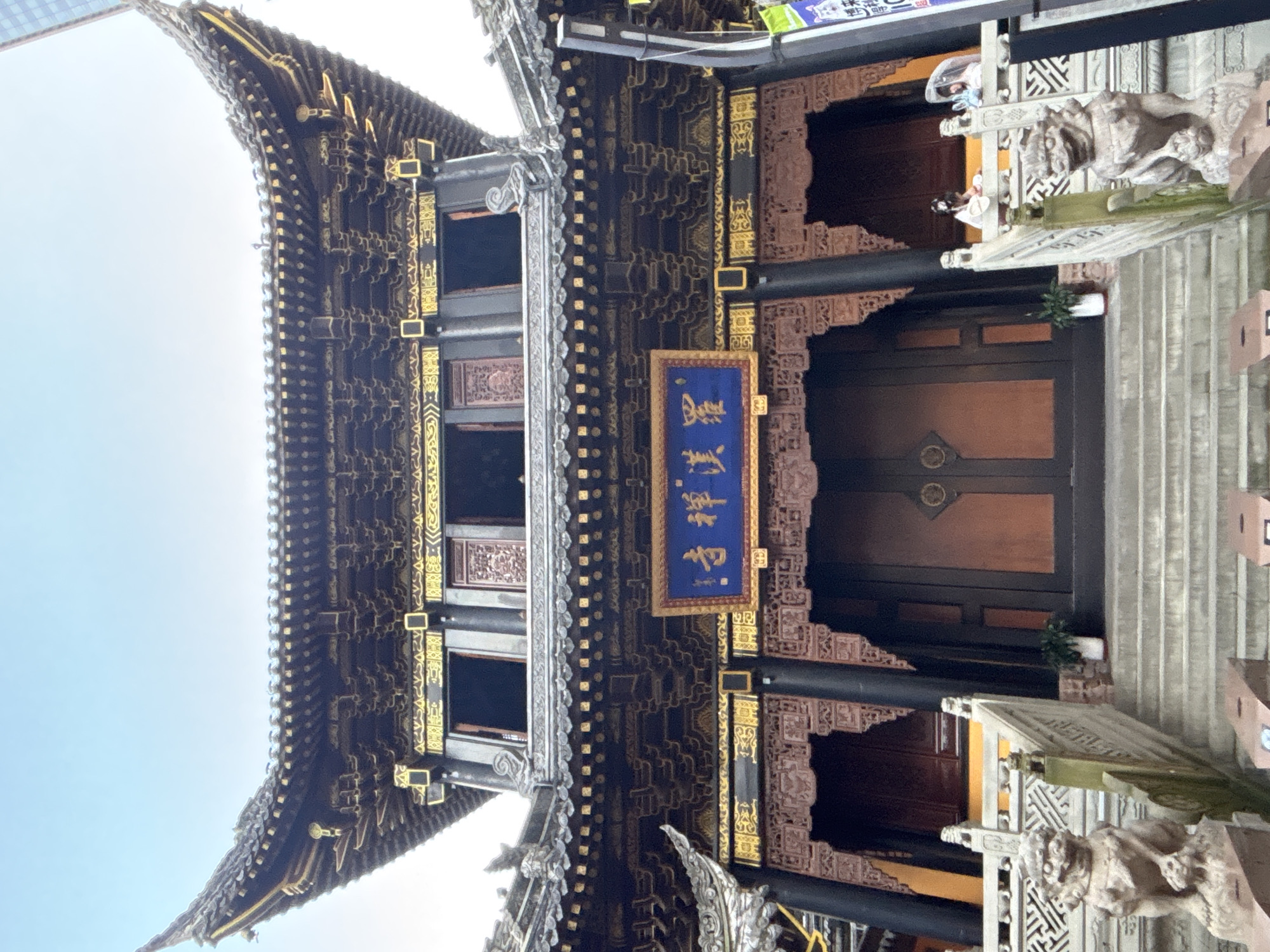 The image shows a close-up, rotated view of the exterior of a traditional Chinese building, likely a temple or historical site, in Yuzhong District, China. The building features intricate dark wooden architecture with ornate gold detailing and decorative carvings. A prominent blue sign with gold Chinese characters is displayed on the facade. The roof has a distinctive curved, tiled design.
Two individuals are partially visible in the background, near a white railing adorned with what appear to be stone animal sculptures, possibly guardian lions. One person is wearing a light-colored top and a face mask, while the other has darker clothing. They are standing on a set of stairs leading up to a large wooden door. A small potted plant is visible near the stairs. The overall atmosphere suggests a daytime scene, possibly with overcast or bright sky, given the lighting. The architectural style and the presence of Chinese text firmly place the location in China. The scene appears calm and offers a glimpse into traditional Chinese craftsmanship and cultural heritage.