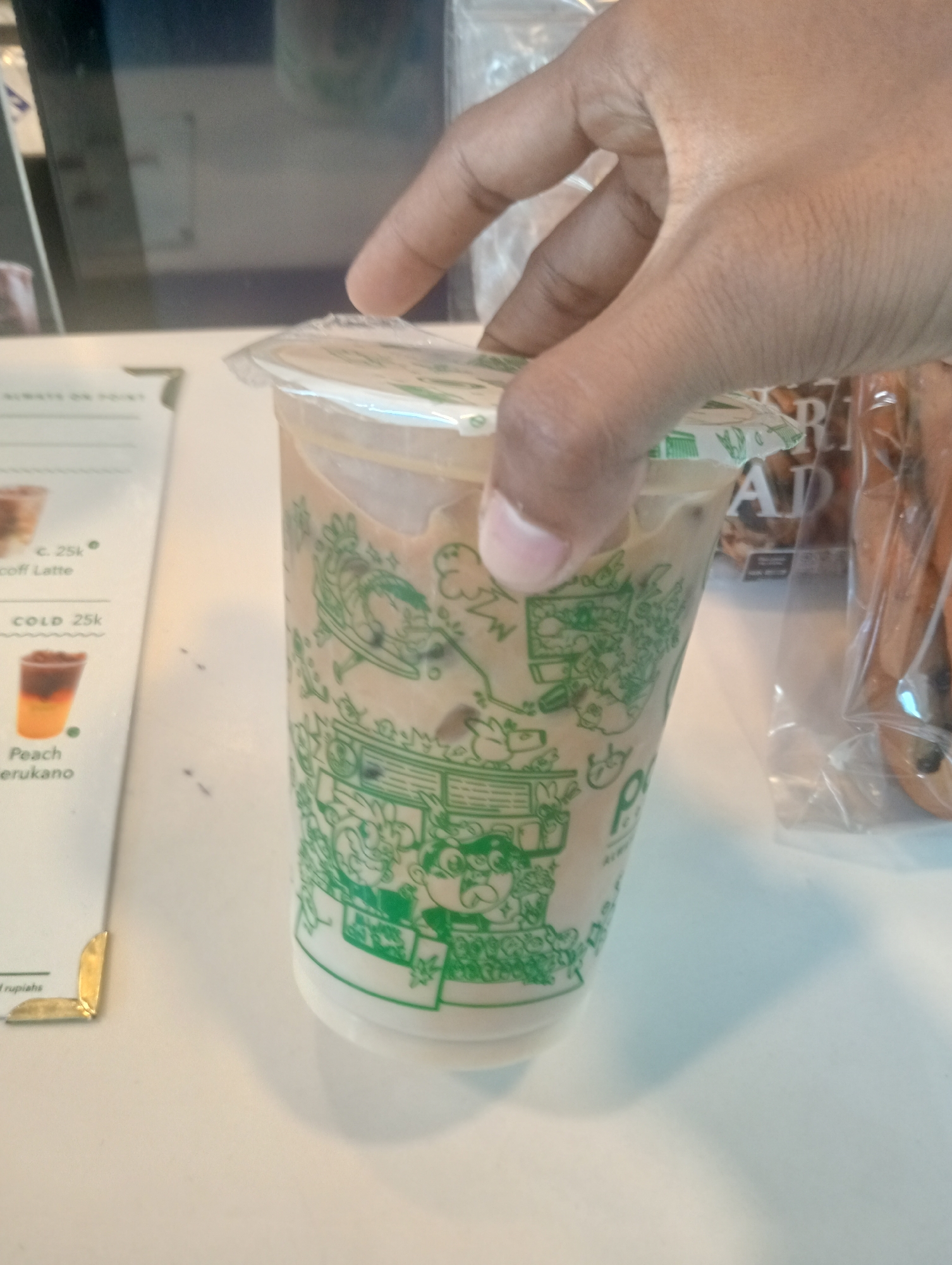 In an indoor setting, possibly a cafe or a retail establishment in Surabaya, Indonesia, a person's hand is holding a clear plastic cup filled with a light brown beverage and ice. The cup is adorned with a green, cartoonish graphic pattern and a clear plastic lid. To the left of the cup is a white menu or information card with some text and an image of a layered drink. In the background, to the right of the cup, is a clear plastic bag containing what appears to be bread or pastries. The scene is set on a white surface, likely a table.