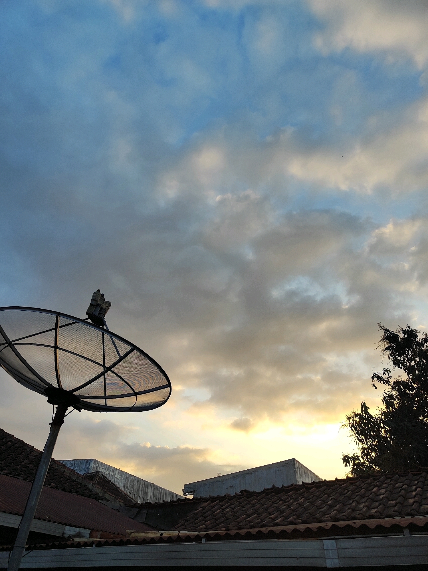 A satellite dish is mounted on a pole above a tiled roof in Kotakulon, Indonesia. The sky above is filled with clouds, with the setting sun casting a warm glow through them. In the foreground, the metallic mesh of the satellite dish is clearly visible, with its supporting structure rising from the roof. The roof itself is made of reddish-brown tiles, and parts of other buildings are visible behind it. A silhouette of a leafy tree branches out from the right side of the frame. The scene depicts a common urban or suburban rooftop view during twilight.