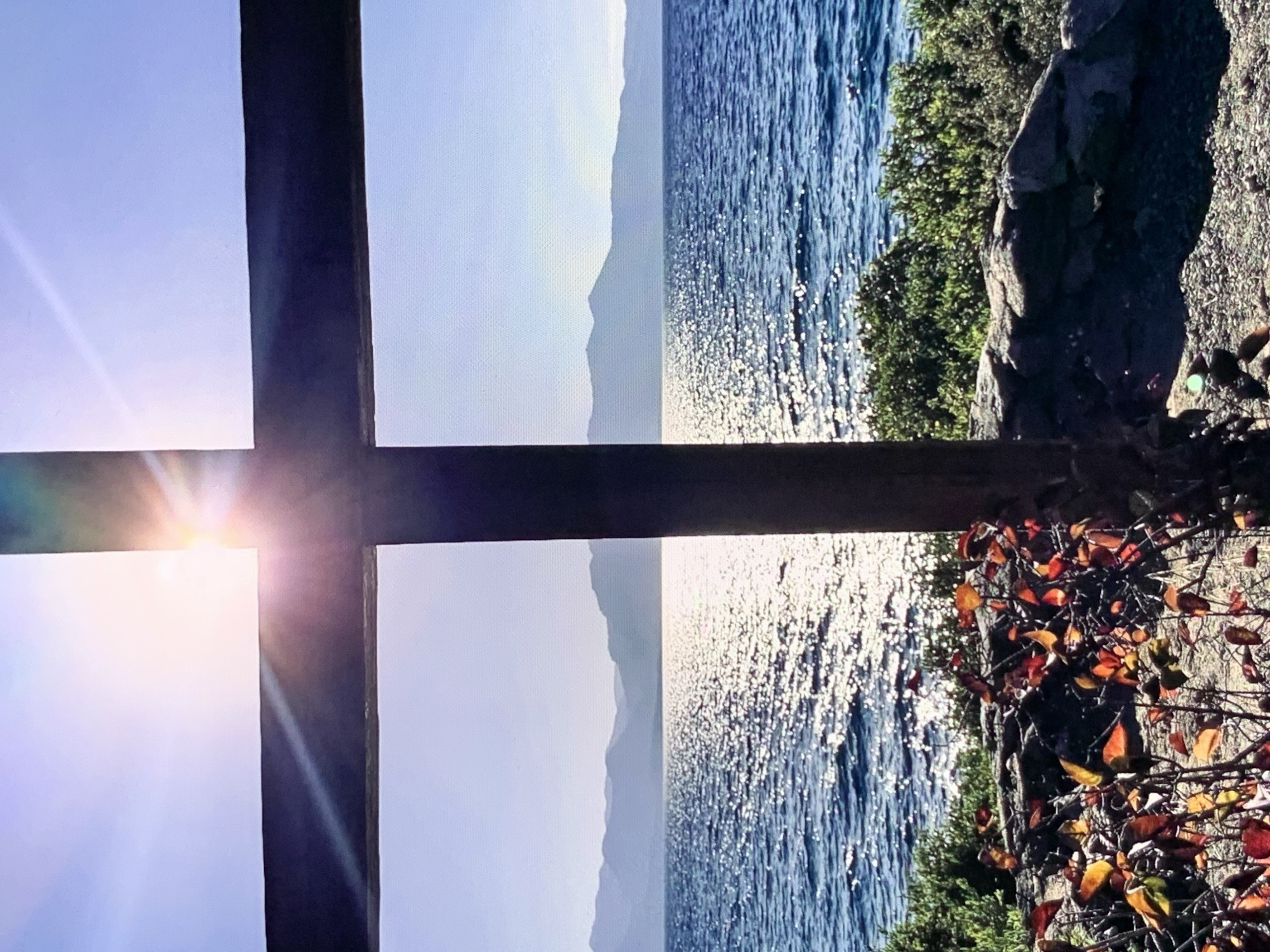 An outdoor, eye-level, vertical, medium shot captures a large wooden cross in silhouette against a bright, sunlit sky and a body of water. The cross is dark brown and positioned in the foreground, occupying the center and right portions of the frame. Sunlight beams through a gap in the cross's upper section, creating a strong lens flare and casting rays across the scene.
Beyond the cross, a vast, sparkling blue body of water stretches to the horizon. The water's surface is textured with ripples and reflects the bright sunlight, creating a shimmering effect. In the distance, a range of layered, hazy mountains forms the backdrop, silhouetted against the pale blue sky.
The immediate foreground, below the water and beside the cross, is composed of rocks and dense green shrubbery. Interspersed among the foliage are several branches adorned with vibrant red and orange leaves, suggesting an autumn setting. The ground in front of the rocks is covered with small pebbles. The scene appears serene and bathed in the warm glow of the sun. There are no people visible in the image. The location is unknown.