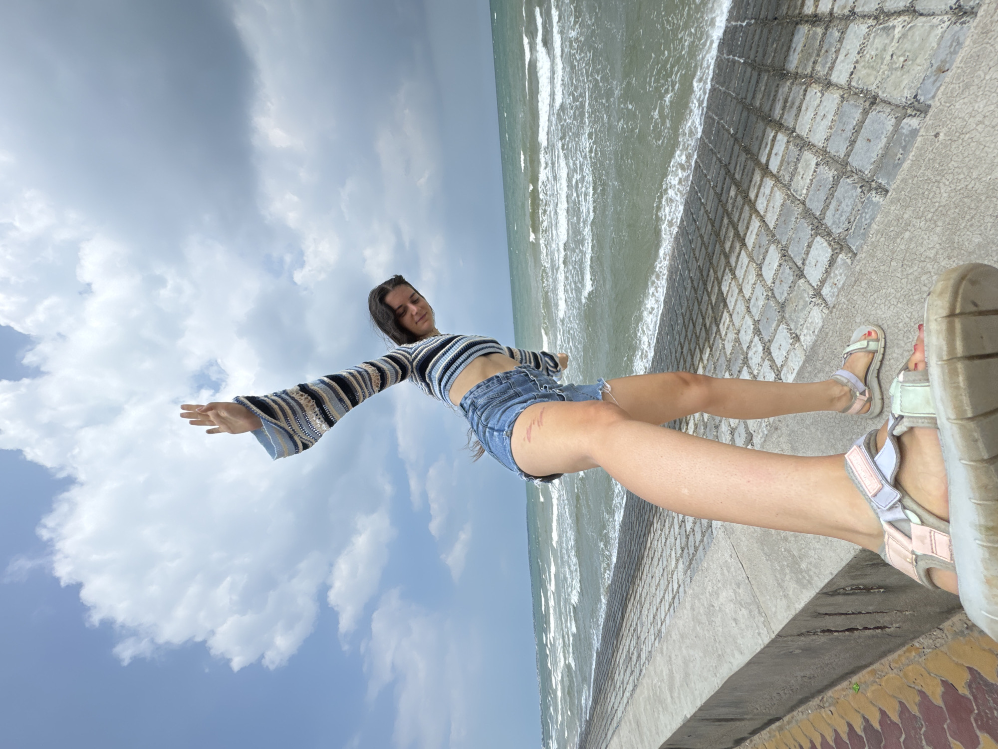 A young woman, with dark hair and wearing a striped long-sleeved crop top and denim shorts, stands on a concrete embankment overlooking the sea. Her left arm is raised above her head, with her hand open, as if waving or in a gesture of freedom. Her right leg is extended forward, and her body is positioned at an angle, suggesting she is either walking or posing for a photograph. The sea is choppy with white waves, and the sky is partly cloudy with a mix of blue sky and dark clouds. The embankment is made of concrete blocks, with a textured pattern, and there is a decorative tiled border on the left side of the image. The scene appears to be a coastal area in Phu Thuy Ward, Vietnam, likely a public promenade or walkway.