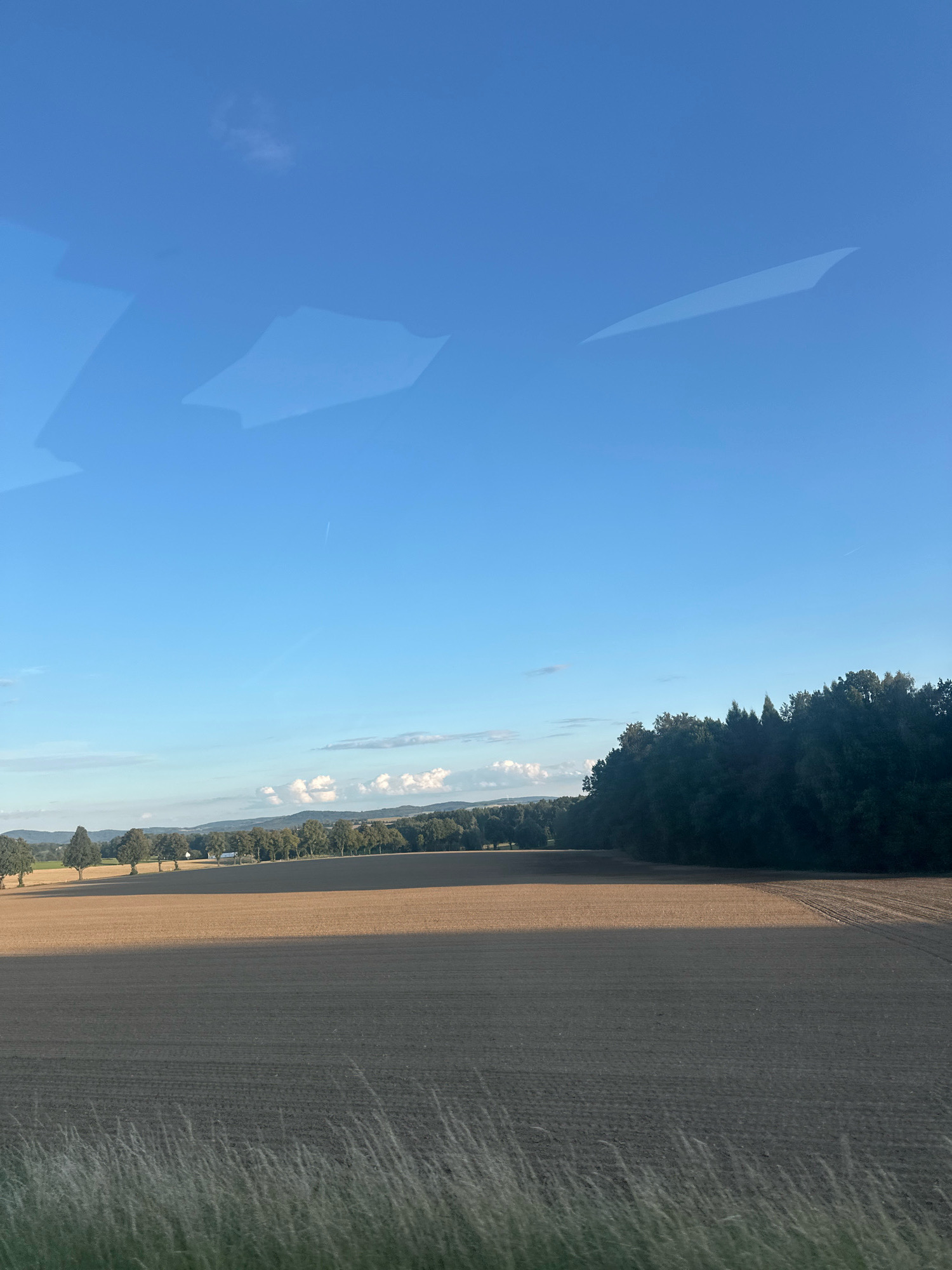 The image depicts a wide, agricultural landscape under a clear blue sky. In the foreground, dry grass is visible. The middle ground is dominated by a large, tilled field, with distinct rows from plowing visible. A sharp shadow divides the field, indicating the direction of sunlight. A treeline runs along the horizon, with a dense forest on the right side of the image and scattered trees and some buildings in the distance to the left. The sky is mostly cloudless, with a few wispy clouds near the horizon and a contrail from an airplane high up. The overall scene suggests a rural setting, possibly in the vicinity of Bretnig-Hauswalde, Germany, characterized by open fields and forested areas.