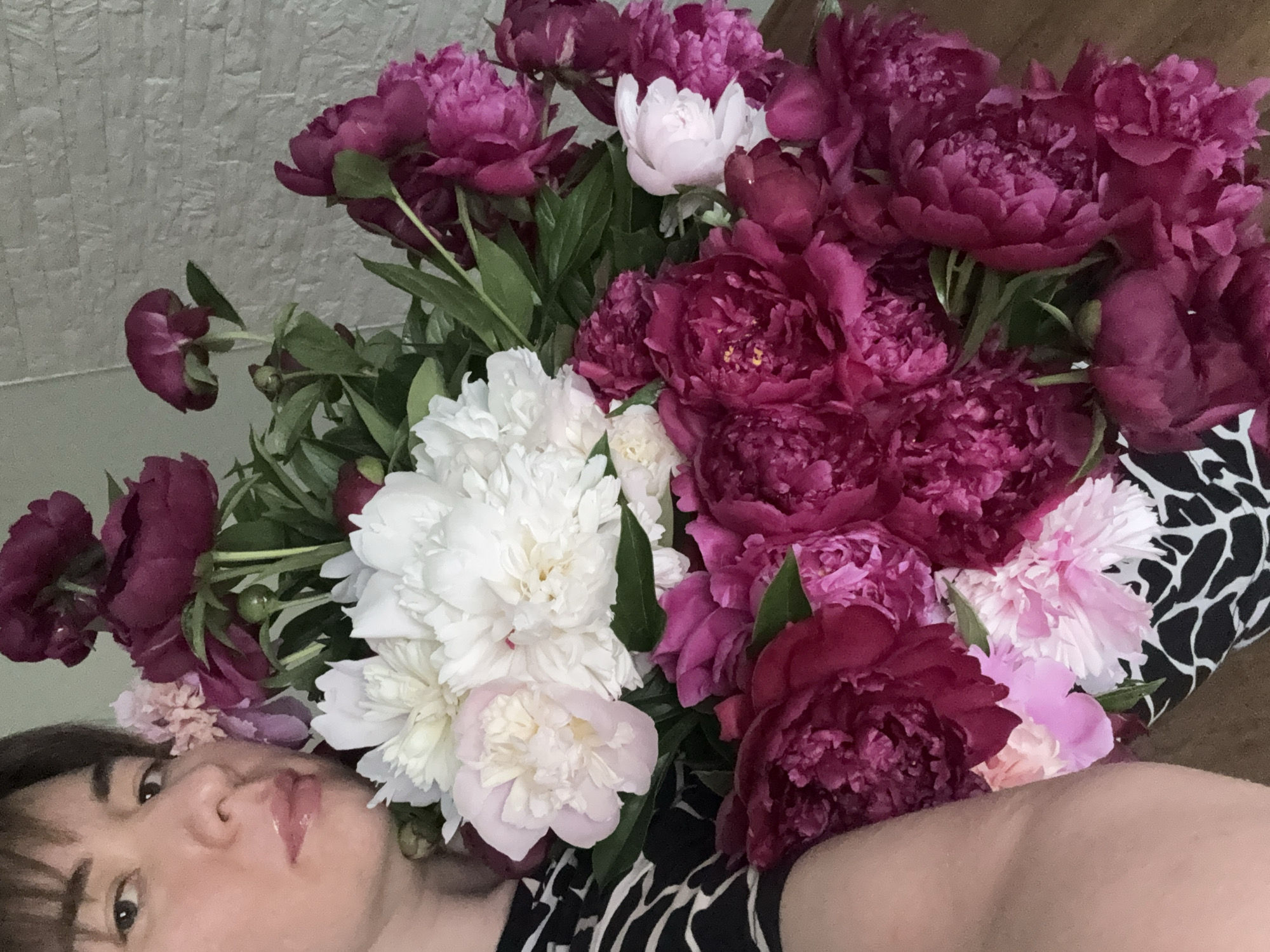 A close-up shot captures a person partially visible in the lower left, positioned in front of a lush bouquet of peonies. The bouquet consists of varying shades of pink, magenta, and white flowers intermingled with green leaves. The person's face is angled towards the camera, with a focus on the eyes and lips. The background appears to be a textured wall, and the person seems to be wearing a black and white patterned garment. The location is Unknown, Unknown.