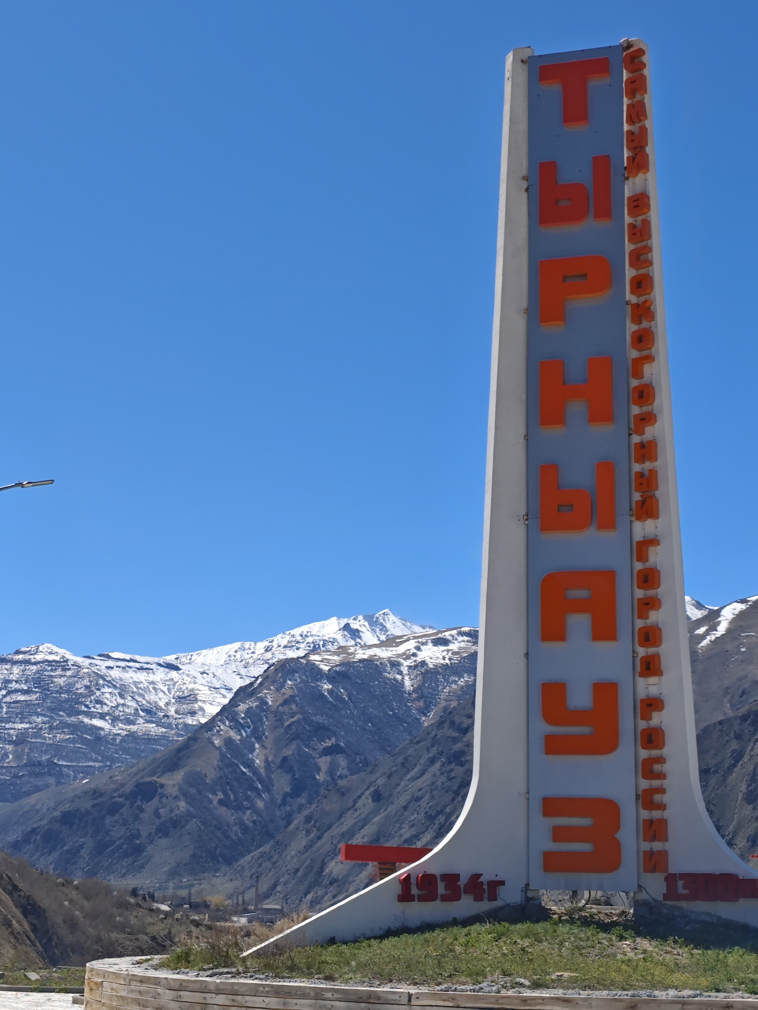 The image shows a tall, white monument-style sign in front of a mountainous landscape with snow-capped peaks under a clear blue sky. The sign is inscribed with the name of the city, "Tyrnyauz" in Cyrillic (ТЫРНЫАУЗ) in large orange letters vertically down the center. Along the right side, there is additional text, also in orange Cyrillic letters that reads "САМЫЙ ВЫСОКОГОРНЫЙ ГОРОД РОССИИ" that translates to "The Highest Mountain City of Russia". At the base of the sign, the year "1934г" is inscribed in orange. The base sits on a wooden-edged platform surrounded by grass. The background features rugged mountains, some covered in snow, and suggests a high-altitude location. There is also what looks to be a road and possibly buildings visible between the mountain and the sign.