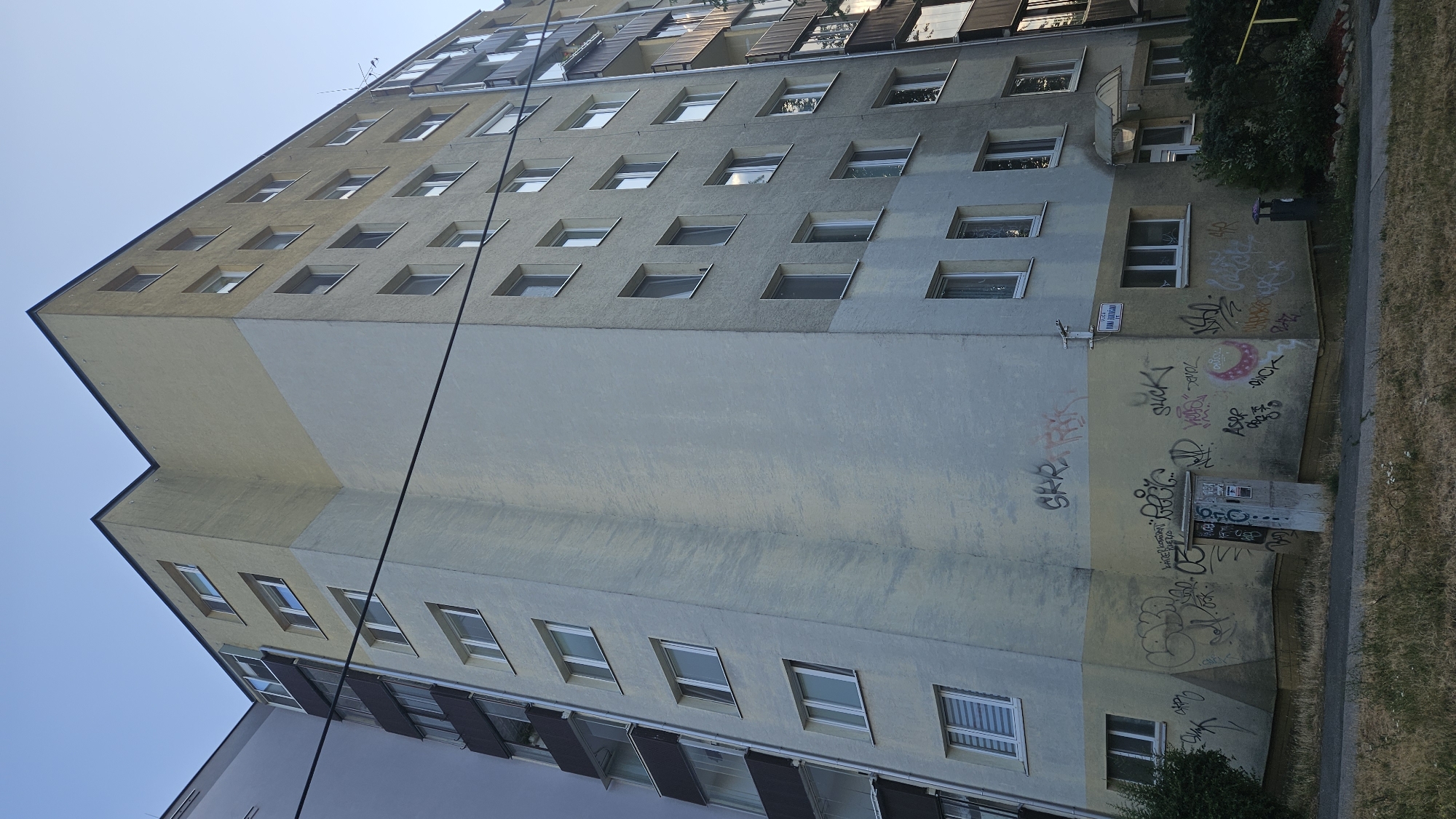 The image shows a multi-story apartment building located on **Jozefova Street in Bratislava, Slovakia**. The building has a distinctive architectural design with two contrasting painted sections, a lower section in a pale yellow and an upper section in a light grey, with a sharp diagonal division between them. The building features numerous windows, many with white frames, and some have balconies with dark railings.
The lower part of the building's facade is heavily covered in graffiti, with various tags and designs spray-painted in multiple colors. A metal utility box is visible near the corner of the building, also adorned with graffiti. A street sign indicating "JOZEFOVA UL." is mounted on the wall.
In front of the building, a patch of dry grass and weeds lines the sidewalk. A paved walkway or road runs along the front of the property. The sky above is clear and blue, suggesting it is daytime, likely late afternoon or early evening given the lighting. The overall scene depicts an urban residential environment, possibly a housing estate typical of some areas in Bratislava.