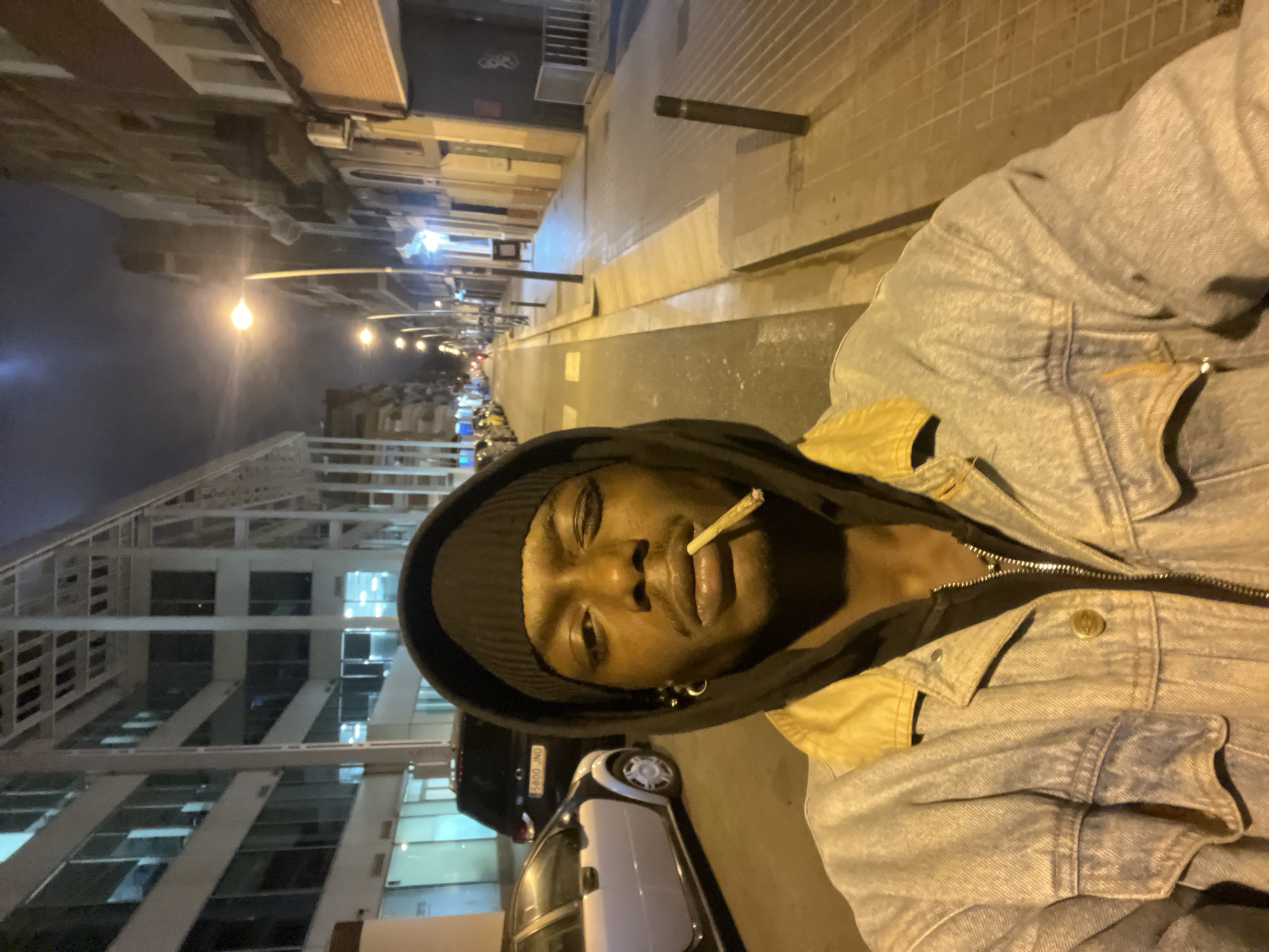 A person with brown skin and short hair is holding a lit cigarette in their mouth. They are wearing a dark beanie, a black hooded shirt, and a faded blue jean jacket with yellow details around the collar. The person is standing on a street with cars parked on the side and buildings of varying heights on either side. Streetlights illuminate the scene, casting a yellowish glow. Based on the architectural style, it is highly likely that the location is in Barcelona, Spain.
