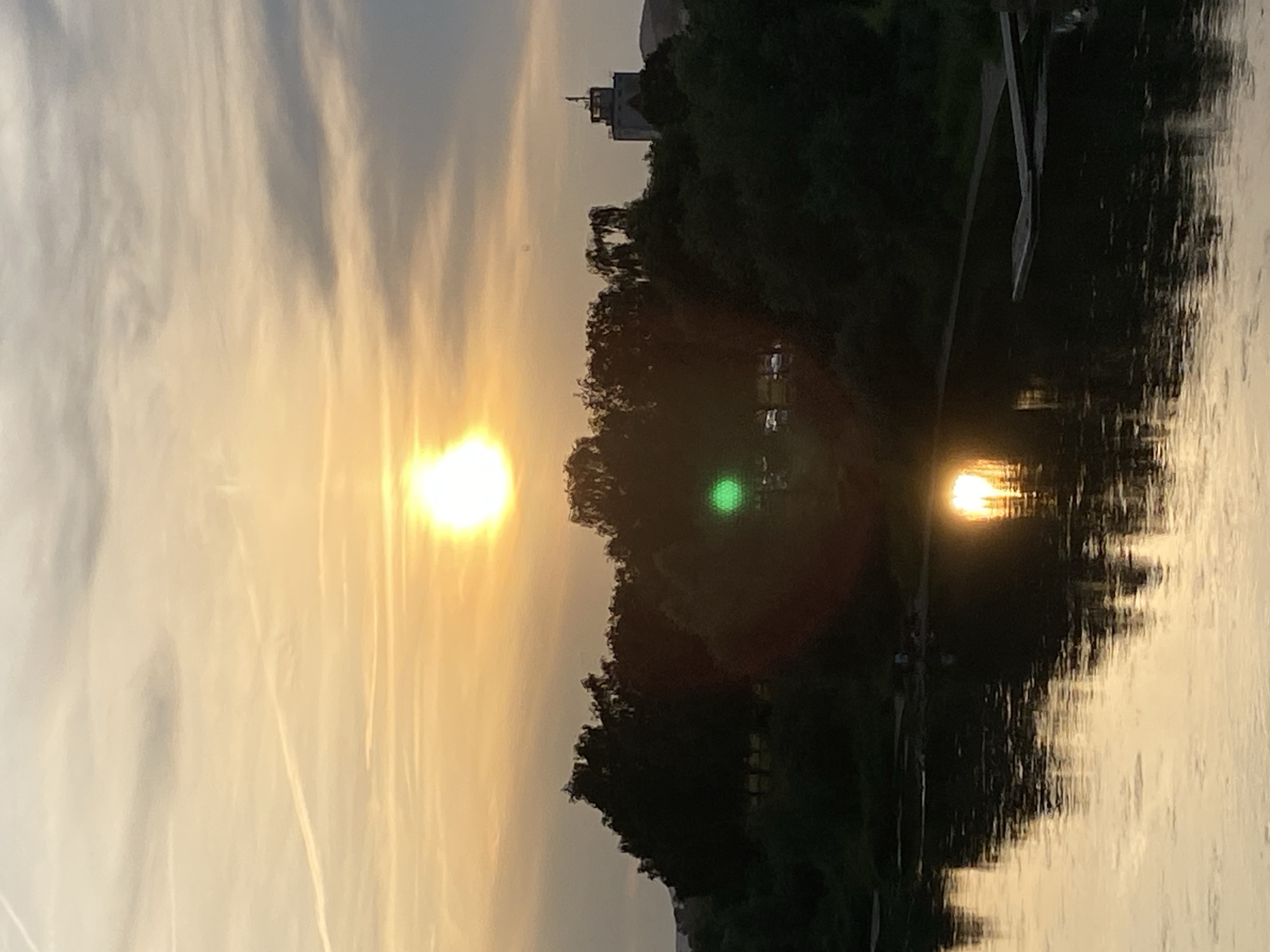 The image depicts a sunset over the Weser River in Rinteln, Germany. The sun, a bright orb, is positioned in the upper center of the frame, casting a warm golden light across the sky and reflecting on the water. A line of trees forms a dark silhouette across the middle ground, separating the river from the sky. In the distance, a tower-like structure is visible above the trees. The water surface is calm, with ripples reflecting the colors of the sunset. There are a few indistinct figures near the riverbank, possibly people enjoying the view or engaging in recreational activities. A wooden structure, possibly a small pier or dock, is visible on the right side of the river. The overall scene is serene and picturesque, capturing the beauty of a sunset over a body of water within a city.