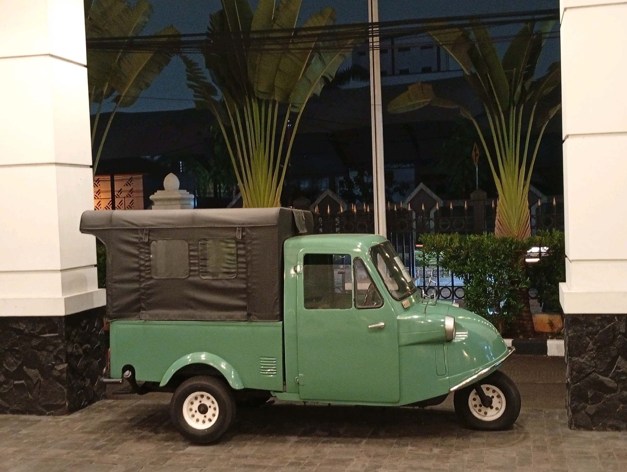 A vintage, green three-wheeled vehicle with a canvas top sits parked on a paved surface. The vehicle has a single front wheel and two rear wheels. The front of the vehicle has a rounded hood and a visible headlight. The canvas top covers the rear cargo area and has two small windows.
The vehicle is positioned in front of a building with large windows and decorative elements. In the background, there are tall, palm-like plants with large fronds. The scene appears to be outdoors, possibly in a courtyard or public space, at dusk or nighttime given the lighting.
The location is identified as Kelapa Dua, Indonesia. The environment suggests a well-maintained outdoor area adjacent to a building. No people are visible in the image. The focus is on the parked vehicle and its surroundings.
