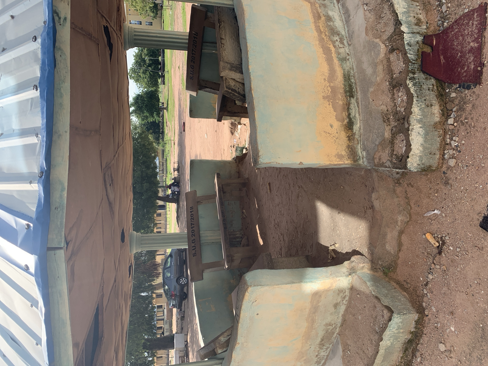 The image depicts an outdoor seating area, likely a gazebo or pavilion, located in Jalingo, Nigeria. The structure features a corrugated metal roof supported by white cylindrical columns. The walls are constructed of concrete and painted a faded light green and yellow, showing signs of wear and tear, with some sections appearing weathered and stained.
Inside the structure, there are two concrete benches with wooden slats for seating. One bench has "S.U.G 2017/2018" engraved on its backrest. The ground within the seating area is covered with reddish-brown dirt and small stones, with some scattered debris including dried leaves and a discarded piece of red material.
In the background, a dark grey sedan is parked, and beyond that, a row of single-story buildings with yellow walls and dark roofs can be seen. Several trees with green foliage are present, suggesting a campus or park-like setting. A few individuals are seated on benches in the distance, partially obscured by trees. The overall impression is of a somewhat neglected but functional public space.