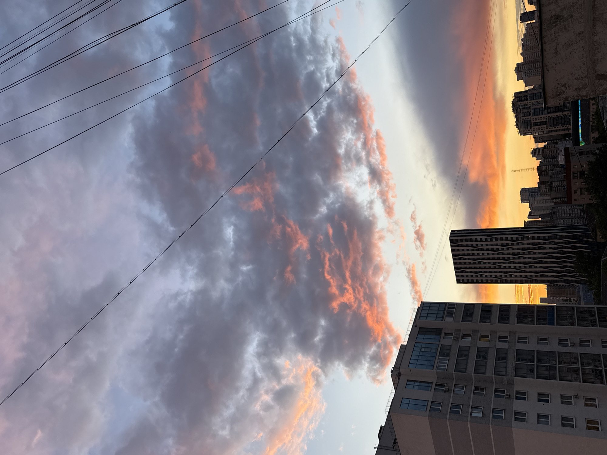 The image shows an upward-angled view of a sunset in an urban environment, specifically the city of Yekaterinburg, Russia. Buildings are visible on the right side, including a tall, modern skyscraper with a distinct black and white pattern. Below the skyscraper, there is a multi-story building with numerous windows. Silhouetted high-rise buildings appear farther in the distance. Overhead, power lines are strung diagonally across the upper left corner of the image. The sky is filled with clouds, some shaded grey, and others tinged with orange and pink from the sunset. The light of the setting sun creates a golden glow in the gaps between the buildings.