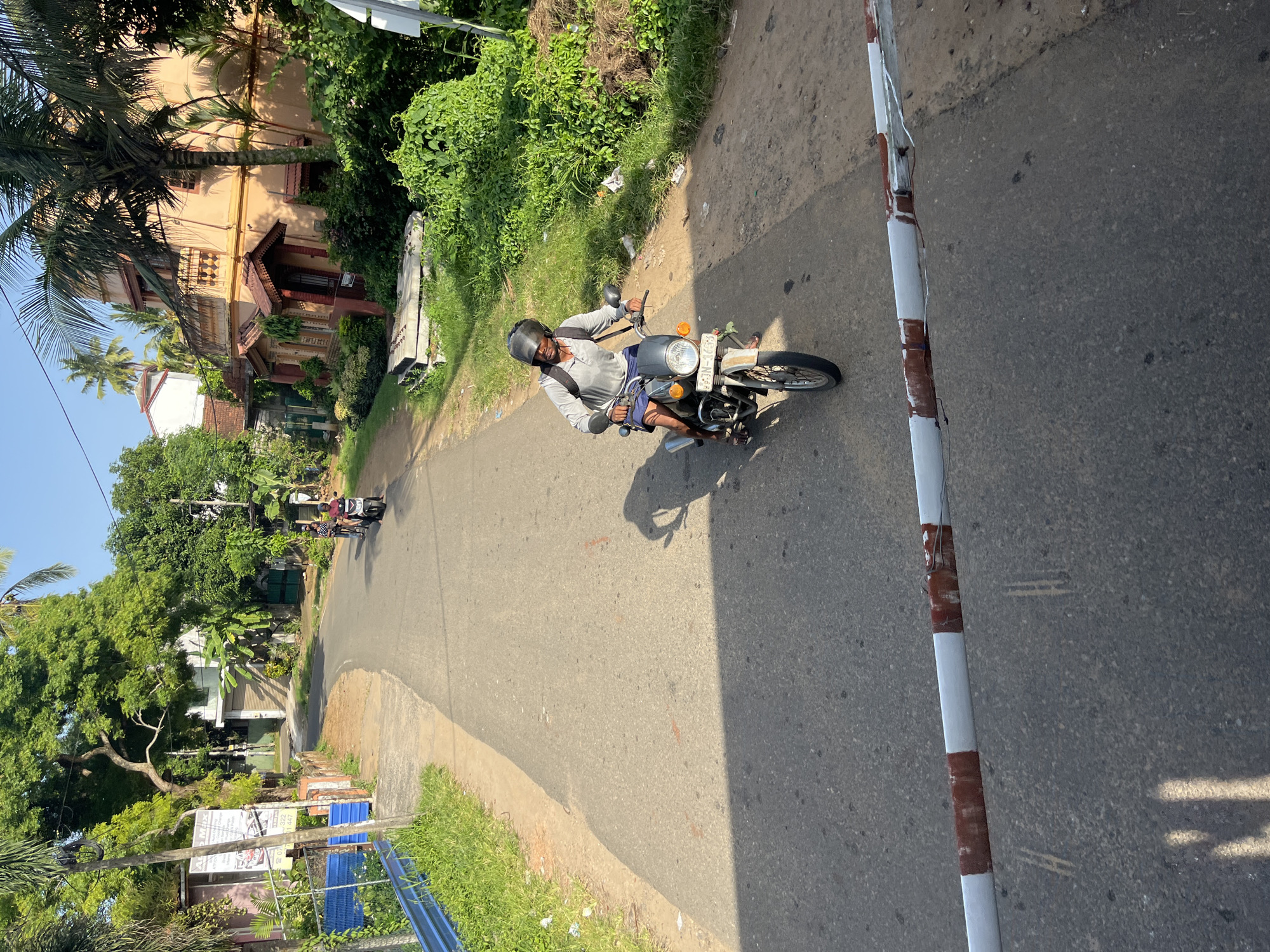 A man rides a motorcycle on a paved road in Kalutara, Sri Lanka. He is wearing a grey long-sleeved shirt, blue shorts, and a black helmet. The motorcycle is a silver and black model with a white license plate. The road is lined with lush green vegetation and trees, with houses visible in the background. The sky is clear and blue. A barrier with red and white stripes is visible in the foreground.