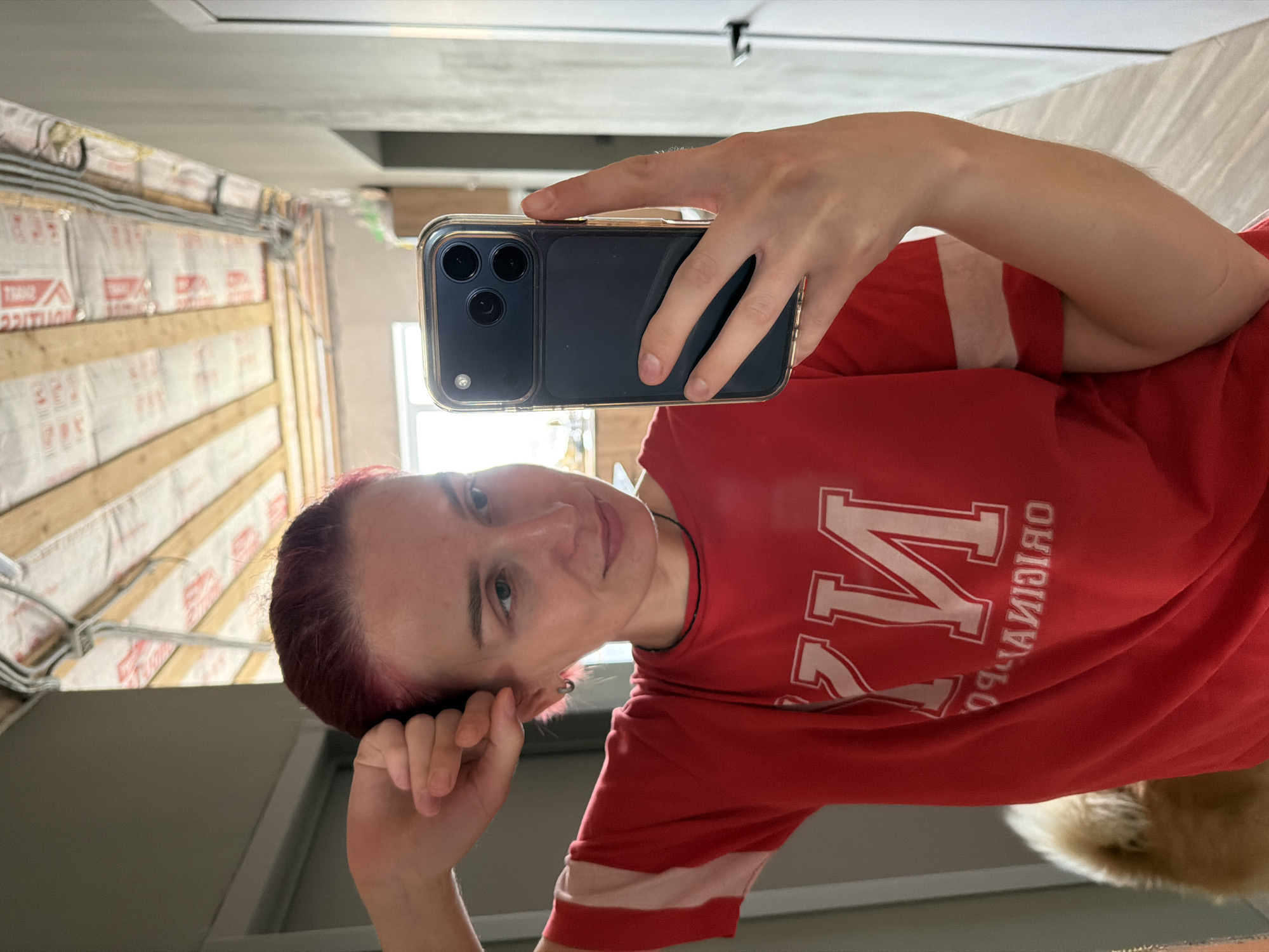 A young woman with short, red hair is taking a mirror selfie in an unfinished room. She is wearing a red t-shirt with white stripes on the sleeves and a white "NY" graphic on the front. Her left arm is raised, touching her hair, and she is looking at her phone, which is held in her right hand. The room has exposed wooden beams and insulation on the ceiling, and unfinished walls. A portion of a golden retriever dog is visible on the left side of the frame.