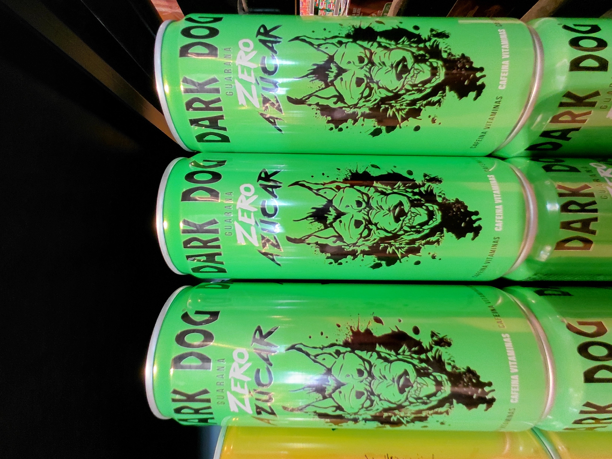 The image shows a stack of three bright green cans of "Dark Dog" energy drink. The cans feature a black graphic of a snarling dog's head. The text "DARK DOG" is prominently displayed in large, dark lettering. Below this, "ZERO AZUCAR" (zero sugar) is written in a stylized font, with "GUARANA" also visible. Smaller text along the side of the cans reads "CAFEINA VITAMINAS". The cans are stacked vertically, with the top can slightly angled. The background is dark, with some hints of wood grain from what appears to be shelving. The lighting suggests an indoor setting, possibly a store or storage area. The overall impression is of a product display.