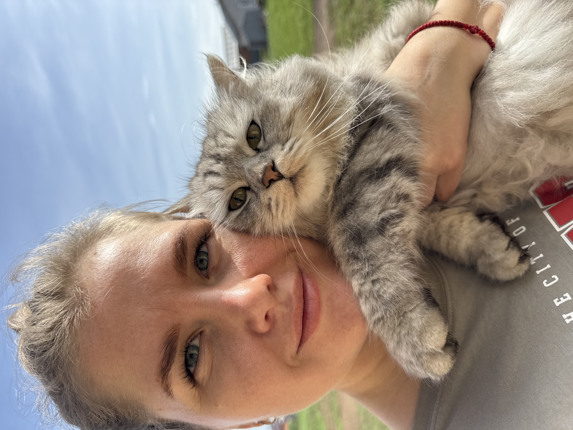 A woman is holding a gray tabby cat against her shoulder in an outdoor setting in Ферма, Russia. The woman has light skin, fair hair pulled back from her face, and blue-green eyes. She is wearing a gray t-shirt and a red bracelet on her wrist. The cat has long fur, green eyes, and a somewhat grumpy expression. The background includes a blue sky, some greenery, and a glimpse of a building. The overall angle of the image is tilted upwards.