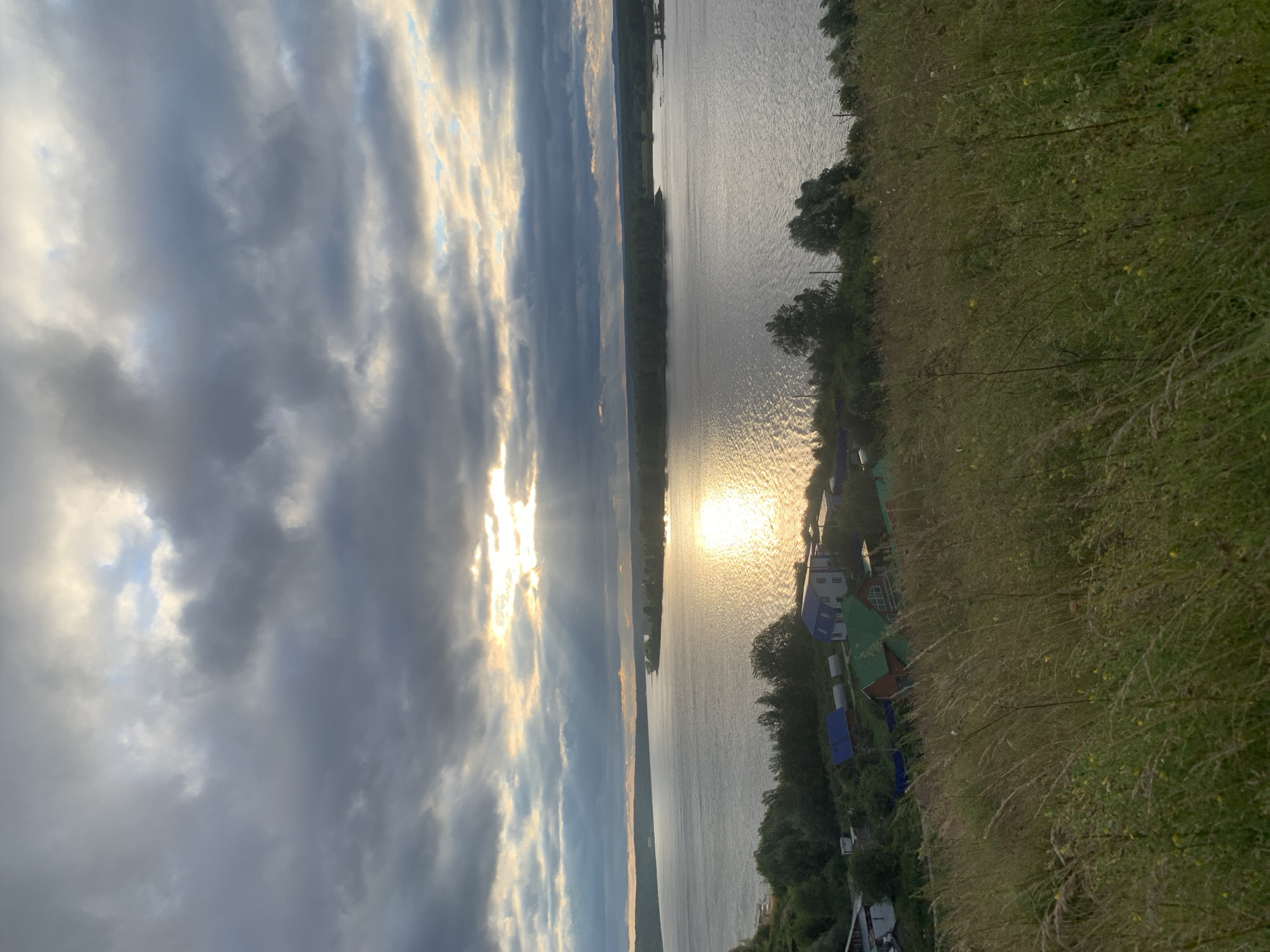 The image depicts a wide river in Olkhovka, Russia, under a dramatic sky filled with dark, heavy clouds. Sunlight pierces through a gap in the clouds, casting a brilliant golden reflection on the water's surface. Along the riverbank, a small cluster of houses with colorful roofs – blue, green, and red – are visible amidst lush green vegetation and trees. The foreground is dominated by a grassy slope covered in tall, wind-blown grasses. The scene suggests a serene, rural landscape with a touch of dramatic weather. There are no people or animals visible in the image.