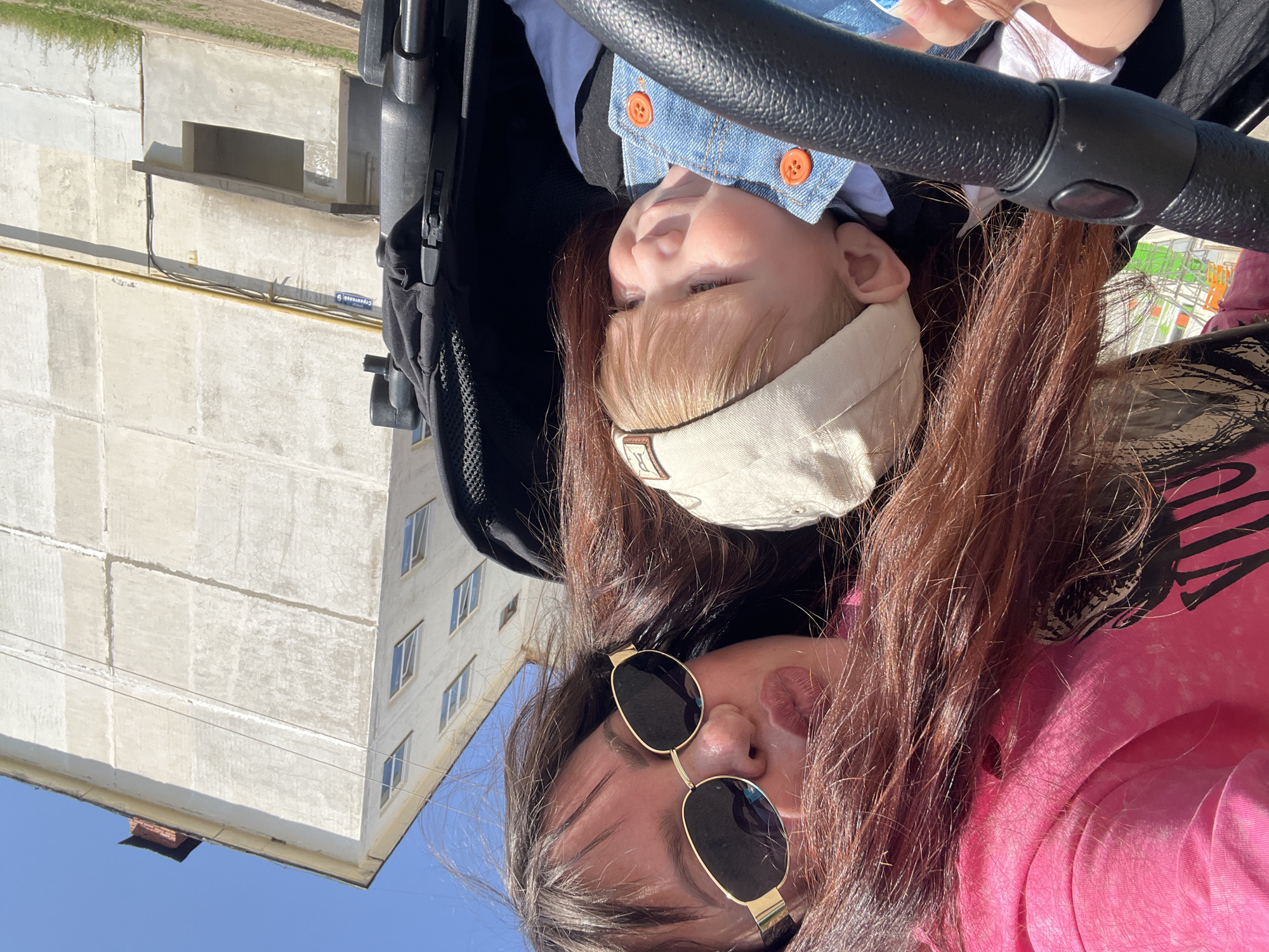 A woman in a pink shirt and gold-rimmed sunglasses is taking a selfie with a baby in a denim jacket and tan cap, both seated in a black stroller. The background shows a light-colored building with multiple windows under a blue sky, and a small portion of green ground cover. The location is Noviy, Russia.