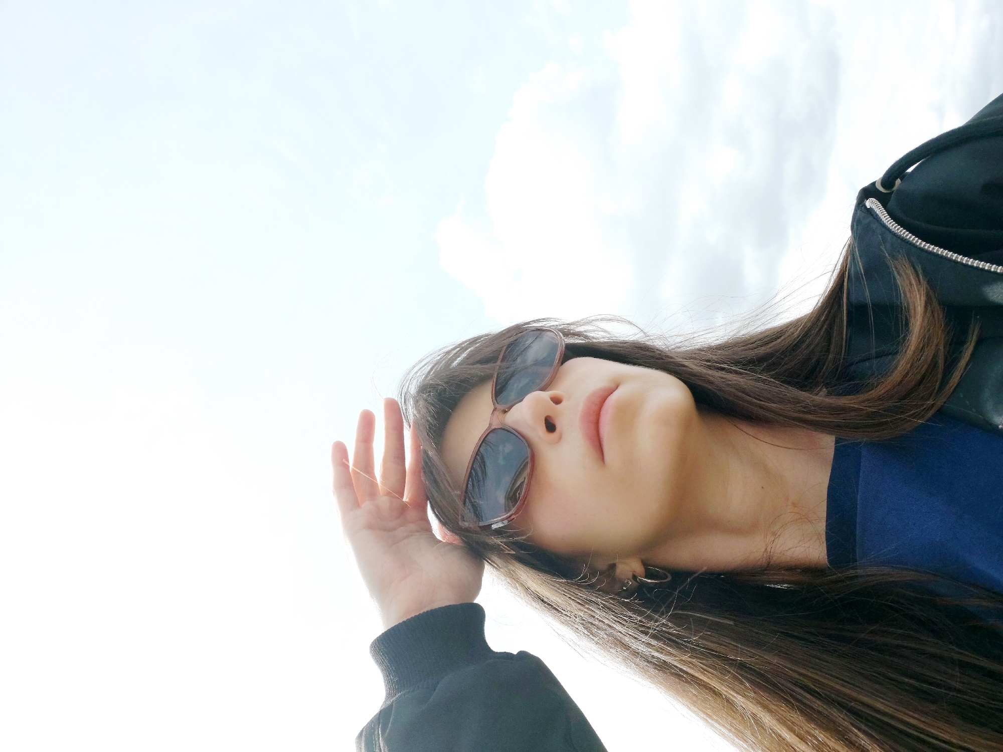 A low-angle shot depicts a young woman with long, dark brown hair wearing sunglasses. She is looking up with her hand partially raised in front of her face. She is wearing a black jacket and a blue shirt. The background is a bright sky with some clouds. There is no visual information in the image to confirm the location as the city of Saint Petersburg, Russia.