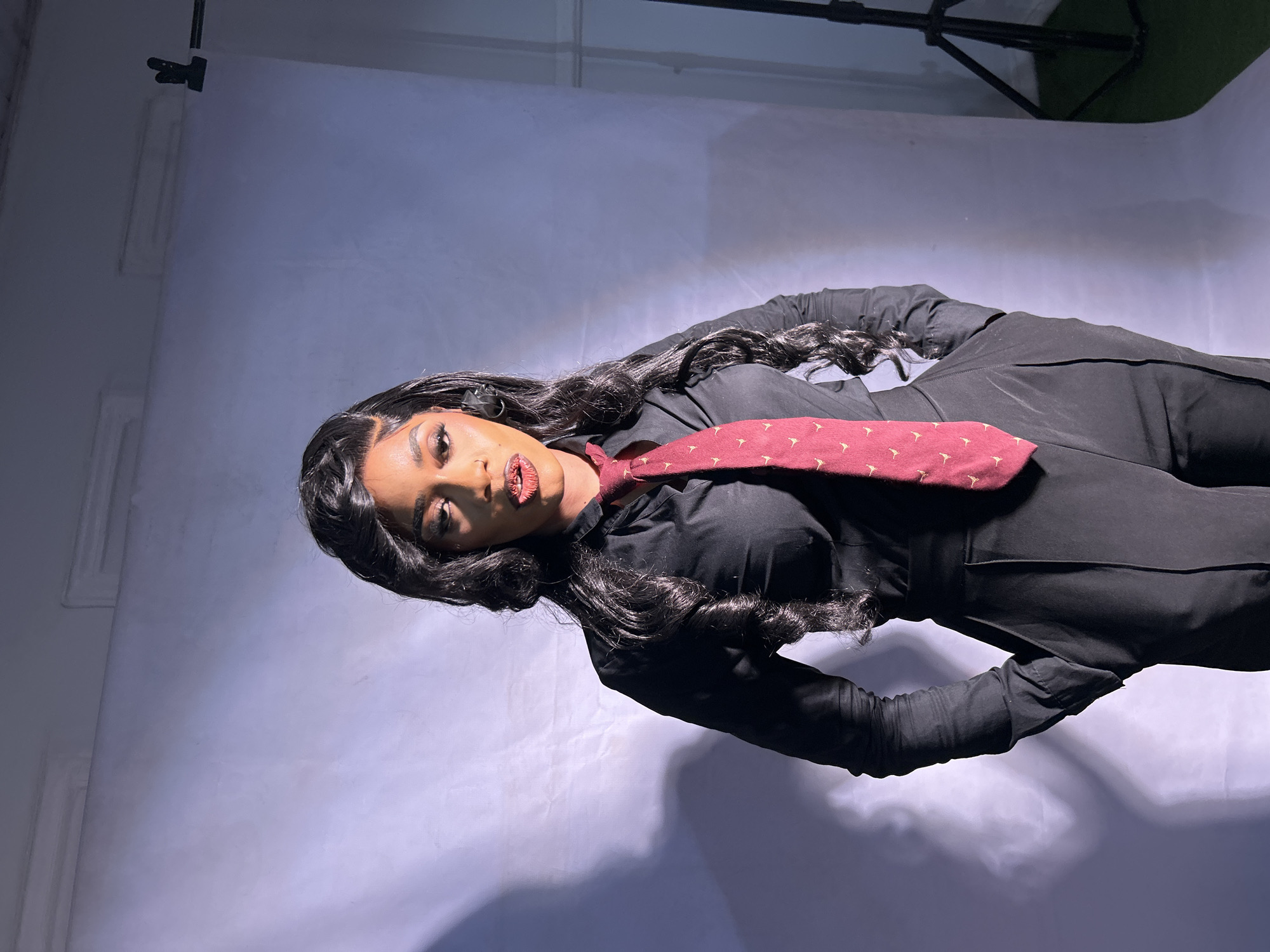 A person with long black hair wearing a black shirt, black pants and a patterned red tie, is photographed against a white backdrop. The image appears to be taken indoors, perhaps in a studio setting. A black object is visible in the background. The location is not discernible from the image.