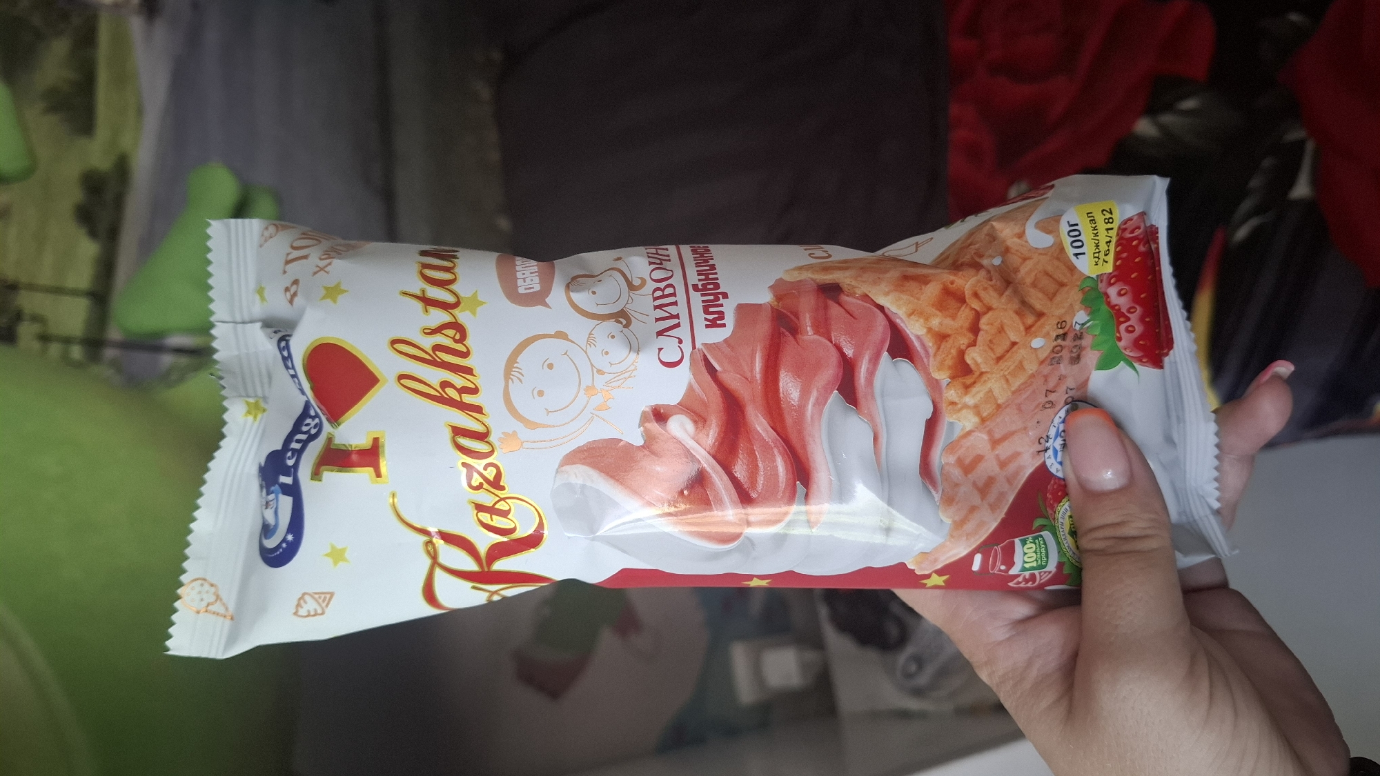 A hand with manicured fingernails is holding an ice cream cone wrapper. The wrapper is predominantly white with red and yellow accents. The prominent text on the wrapper reads "I Kazakhstan" in a stylized font. Below this, there is an image of soft-serve ice cream, with swirling pink and white layers, within a waffle cone. The wrapper also features cartoon drawings of children, strawberries, and a small label indicating "100г кДж/ккал 764/182" and a date "12 07 2016". The background is blurred, showing what appears to be a green plush toy and a dark surface, possibly a bed. There is no discernible information or imagery in the photo that specifically places the scene in the city of Shymkent, Kazakhstan.