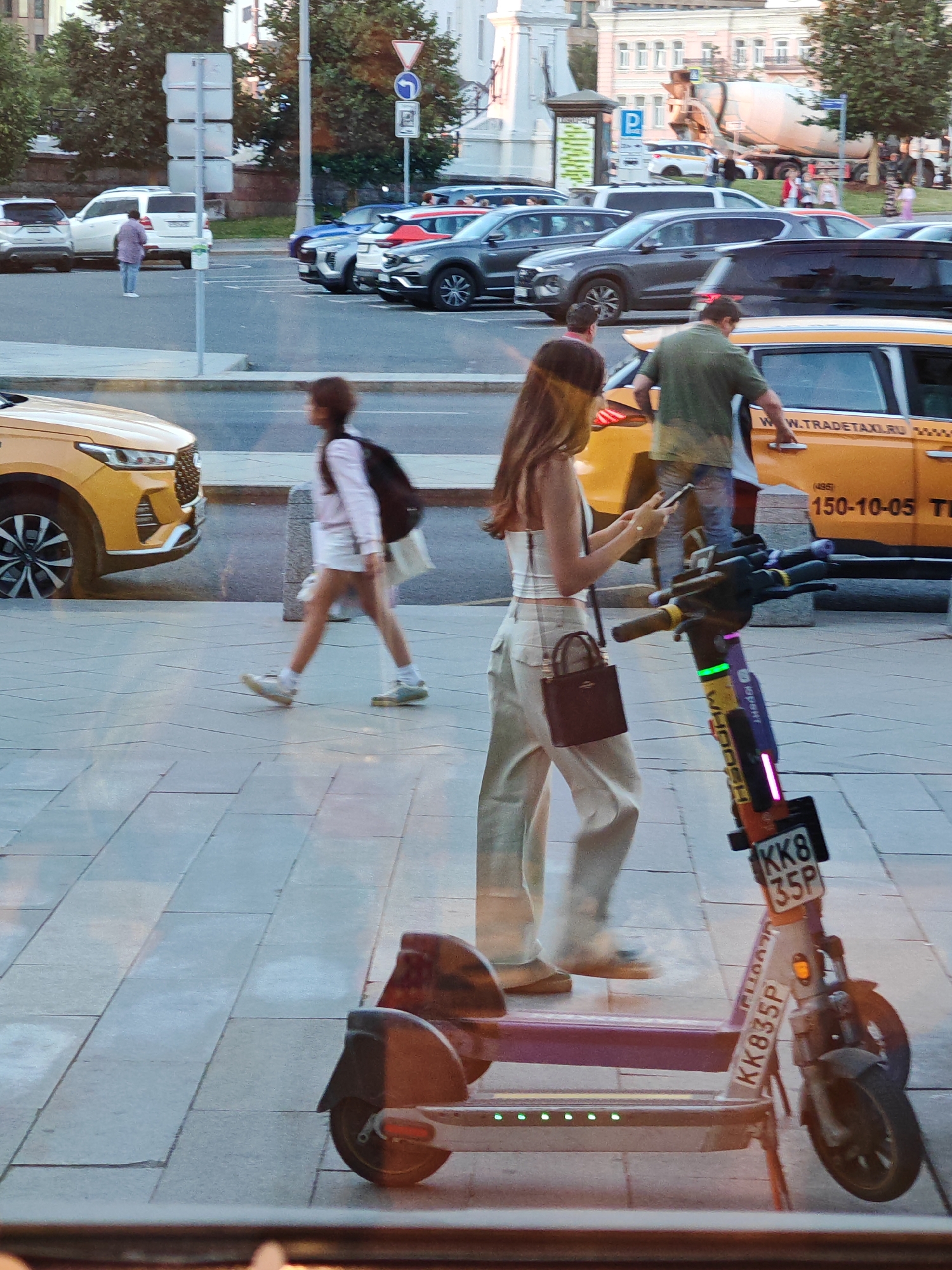 A blurry, reflective shot captures a city scene in Moscow, Russia, likely during the daytime. In the foreground, a purple electric scooter with a license plate reading "KK8 35P" stands prominently. A young woman, partially obscured by reflection, walks past the scooter, holding a phone and wearing a white top, beige pants, and a small dark purse. Her movement is captured in a blur.
Behind her, a yellow taxi with the text "W.TRADETAXI.RU" and the number "150-10-05" is visible. A man in a green shirt is seen near the taxi, possibly interacting with it. Further in the background, various cars, including a white SUV, a gray SUV, and a blue car, are parked or driving on a street. A concrete mixer truck is also visible in the distance, suggesting ongoing construction or road work.
Several traffic signs and streetlights are present, indicating an urban environment. The reflections on the surface suggest the photo might have been taken through a window or a polished surface. The overall impression is one of a busy street in a Russian city.