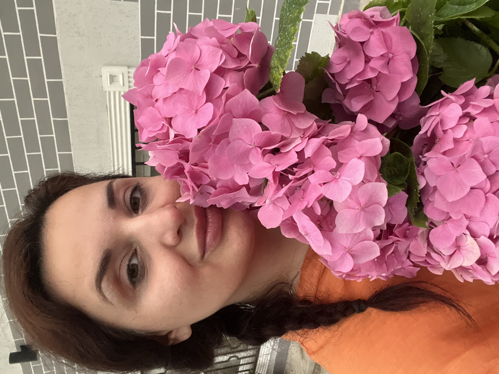 A medium shot shows a woman with dark hair braided over her left shoulder, holding a large bouquet of bright pink hydrangeas. She is wearing an orange shirt. Her head is tilted towards the flowers, and she is looking at the camera with a slight smile. The background consists of a gray brick wall with white horizontal accents. The setting appears to be outdoors, possibly on a balcony or patio.
