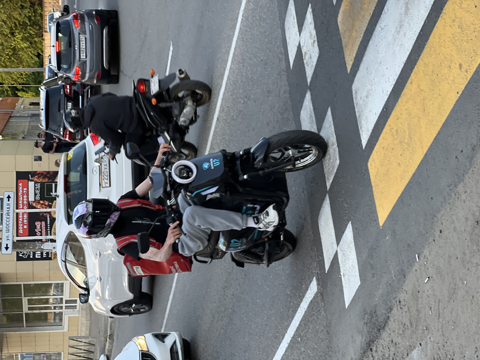 The image shows a street scene in Yablonovskiy, Russia. In the foreground, a person wearing a helmet and a black t-shirt with a red backpack is riding a black electric scooter. The scooter has the text "AIMIFO U2" on its side. The person is wearing grey sweatpants and white athletic shoes. The scooter is crossing a pedestrian crossing with white and yellow markings. In the background, there are several cars, including a white SUV, a black SUV, and a grey SUV. There are also buildings and trees visible. The overall impression is a bustling urban environment on a sunny day.