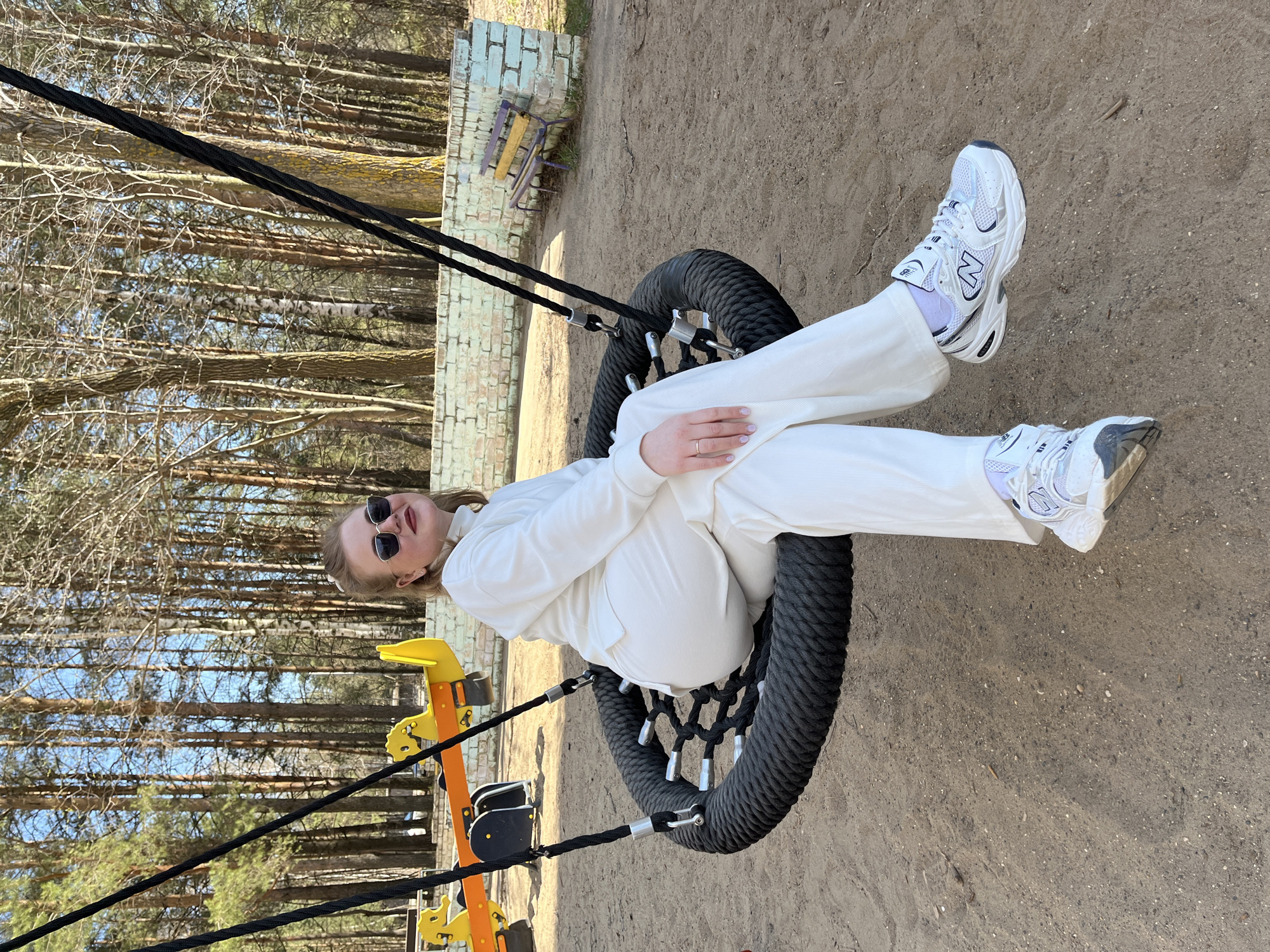 A woman wearing sunglasses and a white tracksuit is sitting in a tire swing at a playground. The swing is suspended from a metal structure with black ropes. The playground is located in Chaikovsky, Russia. The ground is covered in dirt and there are trees in the background.