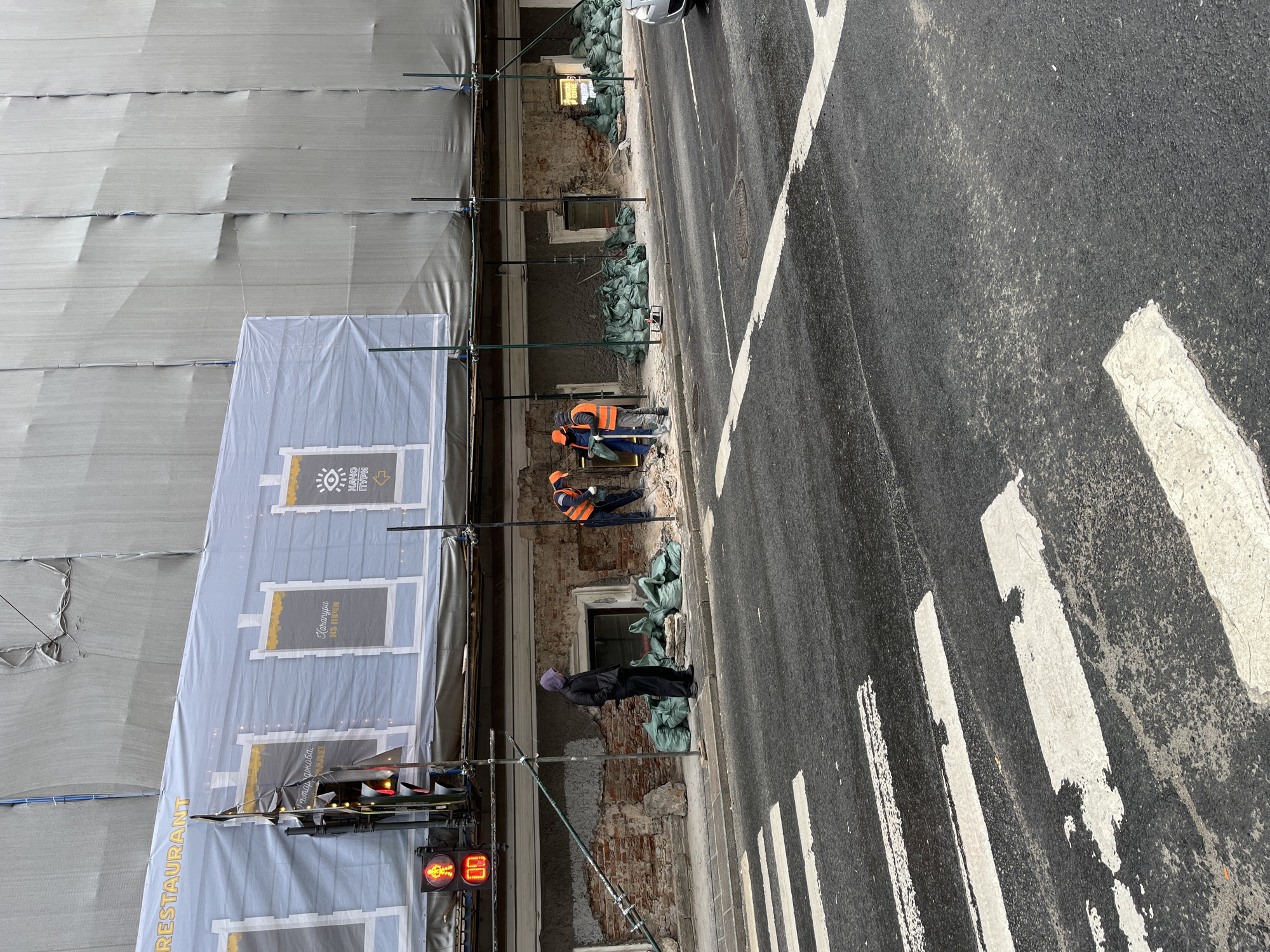 The image presents an urban street scene, oriented vertically, depicting a building under renovation on the left and an asphalt road with a pedestrian crossing on the right. The building's facade is mostly covered by scaffolding draped with plain grey protective tarps. A large banner on the lower part of the scaffolding displays a stylized building design with windows and text. Visible text on the banner includes "RESTAURANT" written vertically, and Cyrillic script such as "МАЙОН", "ОНУМ", and "Барахолка" (flea market), along with an eye icon and an arrow, suggesting a promotional or directional sign. Below the banner, a traffic light shows red signals, including a red pedestrian figure and a red digit, possibly '0' or 'B'.
The lower section of the building reveals old, exposed brickwork interspersed with patches of new concrete or plaster. Numerous green sandbags are piled along the base of the structure. Two construction workers, dressed in orange safety vests and dark pants, are actively engaged in work on the building's facade. A third person, wearing a dark coat and a purple head covering, stands on the sidewalk observing the construction activity.
The asphalt road features faded white pedestrian crossing stripes. Light-colored debris or dust is scattered across the road surface, and a manhole cover is visible further down the street. The lighting indicates daytime, possibly under an overcast sky. The scene captures ongoing renovation work in a public urban setting.