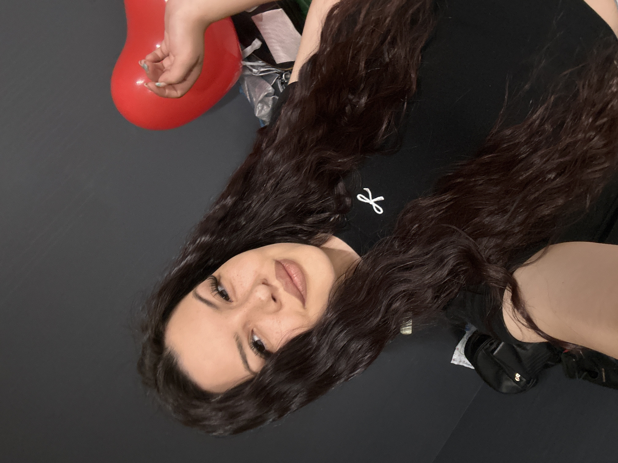 A young woman with long, dark brown, wavy hair is lying on a dark gray surface, possibly a floor. She is wearing a black shirt with a white logo near the collar. Her face is visible, showing light skin, dark eyebrows, and neutral-colored lips. To the left of her head is a red heart-shaped balloon, and partially visible behind her is some plastic wrap. To her right is a black purse. There is no explicit indication of the location being Sliven, Bulgaria.