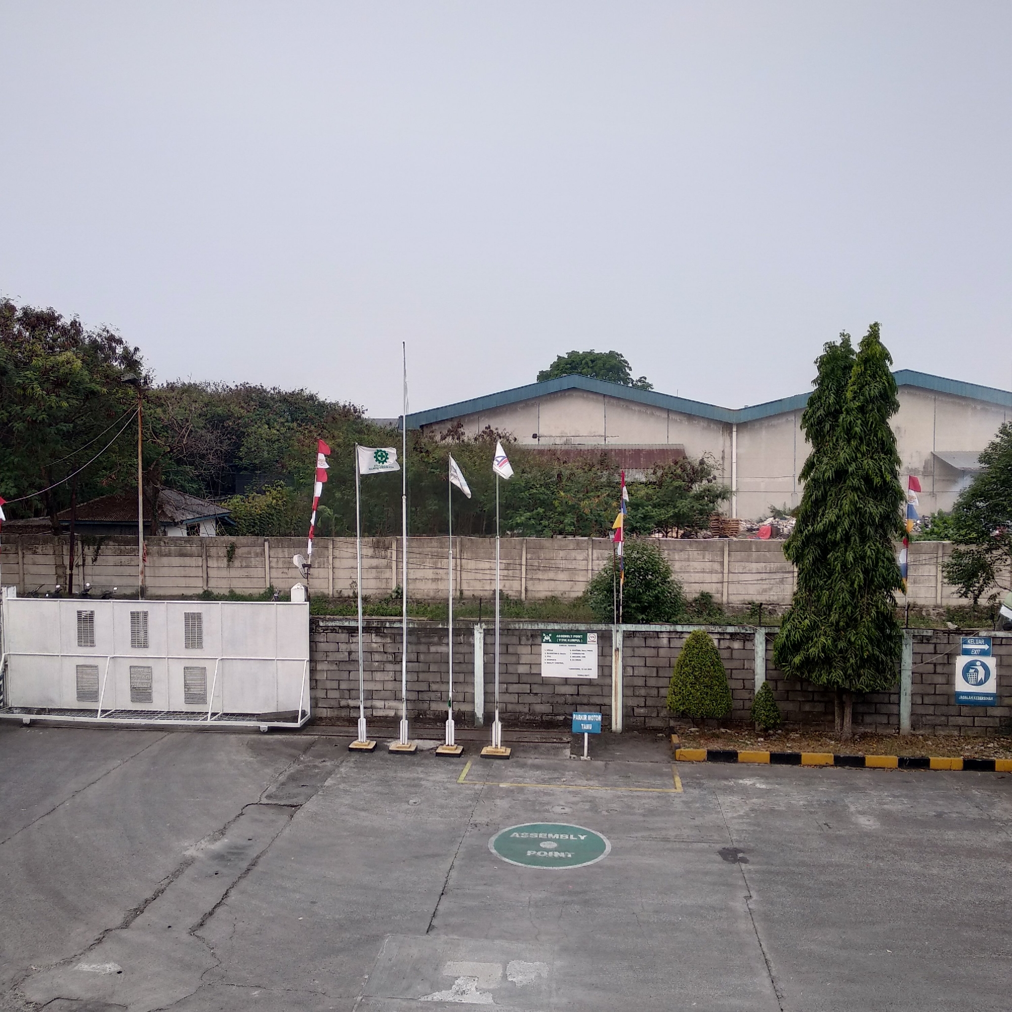 The image shows an outdoor scene at a facility, likely an industrial complex, in Balaraja, Indonesia. The foreground consists of a cracked asphalt surface with yellow and black painted borders along a low brick wall. A white gated structure with multiple ventilation grilles is visible on the left. In the mid-ground, several flagpoles stand, some flying flags, including Indonesian flags and a white flag with a green logo. A concrete wall runs horizontally across the scene, behind which are trees and vegetation. Beyond the wall, a large, light-colored industrial building with a blue roof is visible. Signs are present, including one that reads "ASSEMBLY POINT (TITIK KUMPUL)" and another with a trash bin symbol and the word "KELUAR EXIT" and "JAGALAH KEBERSIHAN". Several trees, including a tall, slender evergreen tree, are interspersed throughout the scene. The sky is overcast and light gray. There are no people or animals visible in the image.