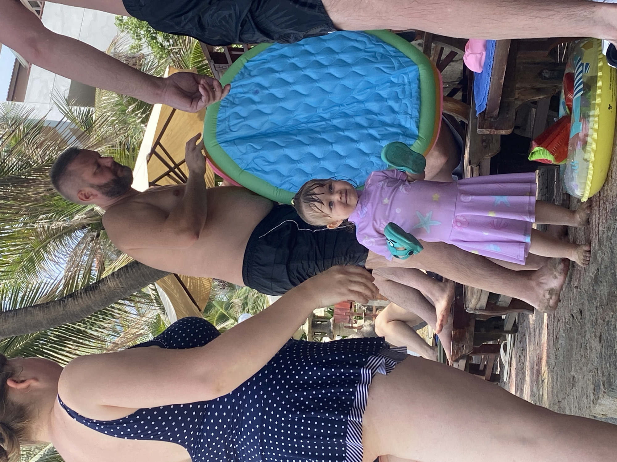 In Phu Thuy Ward, Vietnam, a group of people are gathered near an inflatable pool under palm trees. A woman in a blue polka dot swimsuit stands on the left. In the center, a man with a beard is partially visible, holding a large blue and green inflatable toy with the help of another person whose arm is visible on the right. A young child in a purple swimsuit with a skirt is in the foreground, holding green water shoes. In the background, another person's legs are visible, resting on a wooden bench. The ground appears to be a mix of sand and rough stone.