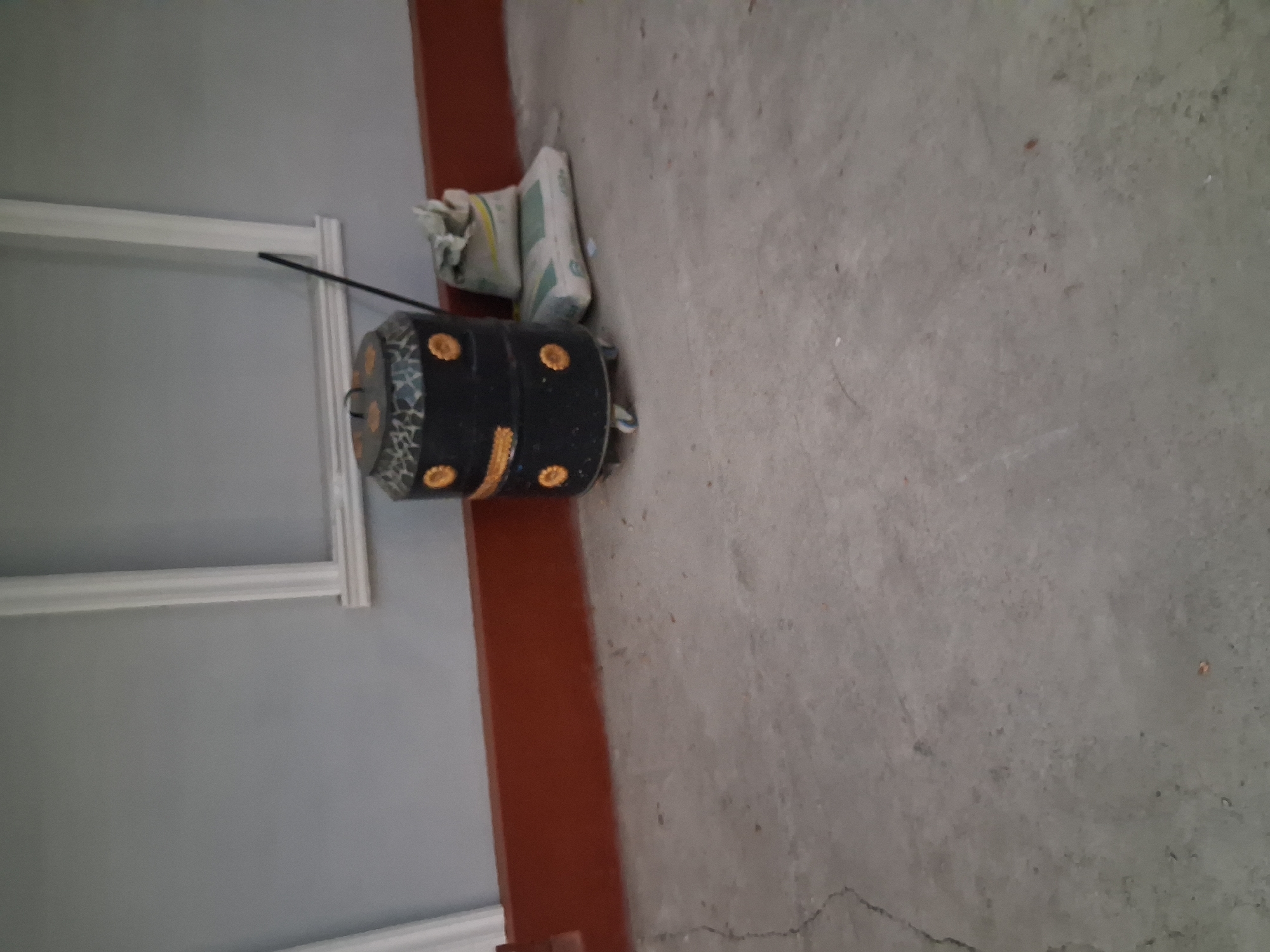 The image shows a close-up, low-angle view of a concrete floor in what appears to be an unfinished interior space. The floor is a neutral gray with some visible cracks and imperfections. To the left, a section of a pale gray wall is visible, with white trim at the bottom and top, suggesting a doorway or window frame that has not yet been fully finished.
In the center-left of the frame, resting on the floor, is a black cylindrical object, possibly a drum or a large container, decorated with a mosaic pattern of blue, white, and yellow tiles. It also has several circular yellow embellishments. Leaning against the wall above and slightly behind this object are two bags of white material, likely cement or similar building supplies, partially covered by a light-colored fabric. A thin black rod or stick extends from behind the decorated cylinder towards the wall.
The scene is lit by an unseen light source, casting minimal shadows and indicating an indoor setting, possibly during daylight hours though the time of day is not clearly discernible. The overall impression is one of a construction or renovation site. No people are visible in the image. There is no visible text. The location context is Kahramon, Uzbekistan.
