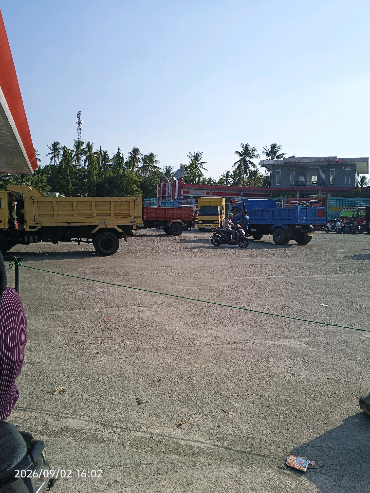 The image shows a bright, sunny day at what appears to be a gas station or a truck depot in Pinrang, Indonesia. The foreground is dominated by a concrete surface with a green rope laid across it. To the left, a portion of a red and white awning is visible, likely belonging to a gas station.
In the middle ground, several trucks are parked. A large yellow dump truck is prominent on the left, with its bed raised. Behind it, a red dump truck is visible. Further to the right, a bright blue dump truck is parked, with two men standing near it. One man is sitting on a motorcycle, facing the blue truck, and the other man stands beside him. Other vehicles, including scooters, are parked in the background.
The background features a line of palm trees and other vegetation, with a tall telecommunications tower visible in the distance. A multi-story building with a modern architectural style, possibly an office or commercial building, is also present in the background, partially obscured by trees. The sky is clear and blue. The timestamp "2026/09/02 16:02" is superimposed on the bottom left of the image.