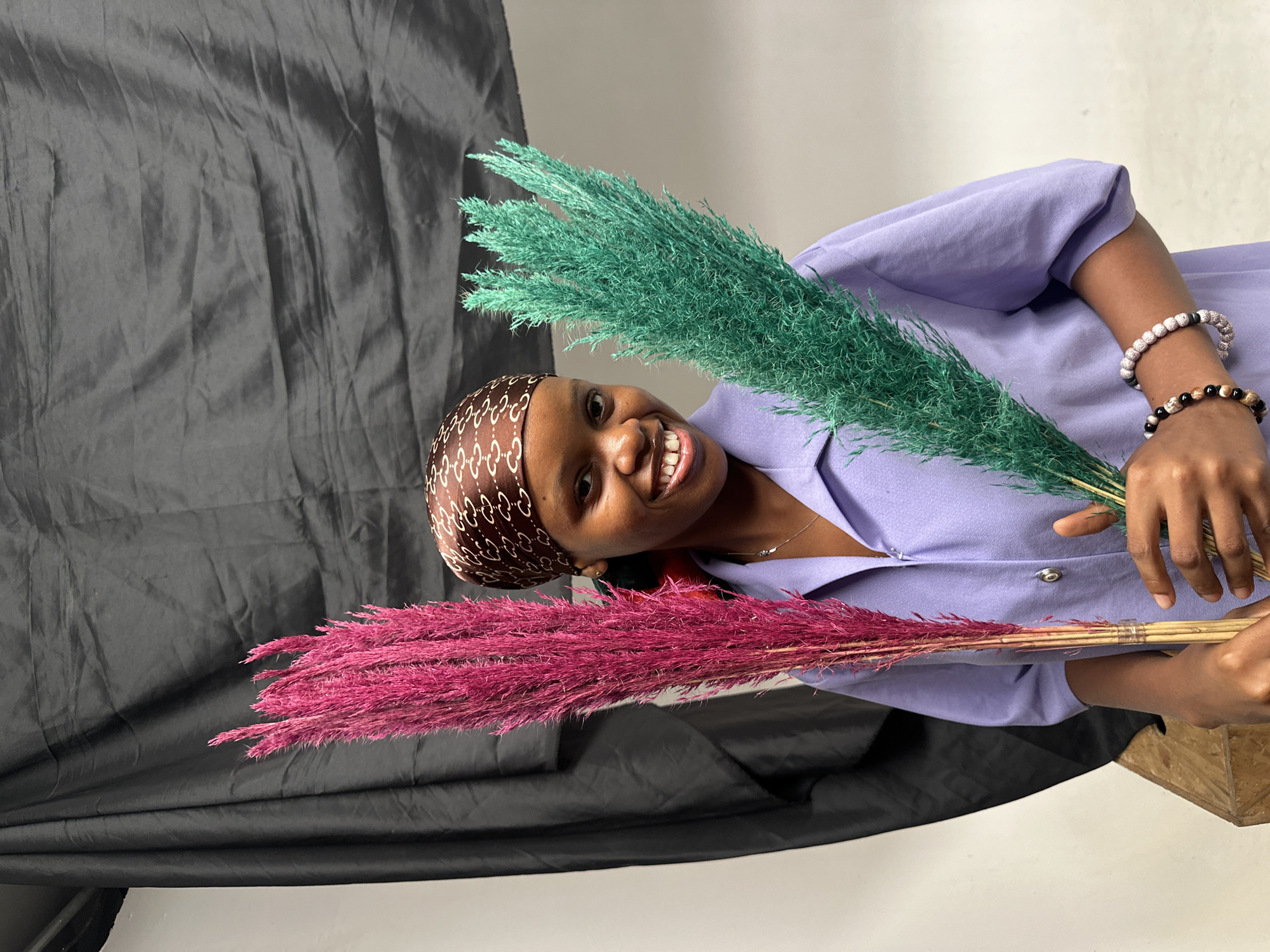 A smiling young Black woman is the primary subject of this indoor photograph. She is wearing a lavender blouse, a brown head covering with a white pattern, and bracelets on her right wrist. She is holding two bunches of dyed reed grass: one green and one magenta. Behind her is a draped black fabric backdrop. The location is likely a studio or an indoor space, possibly in Ikeja, Nigeria.