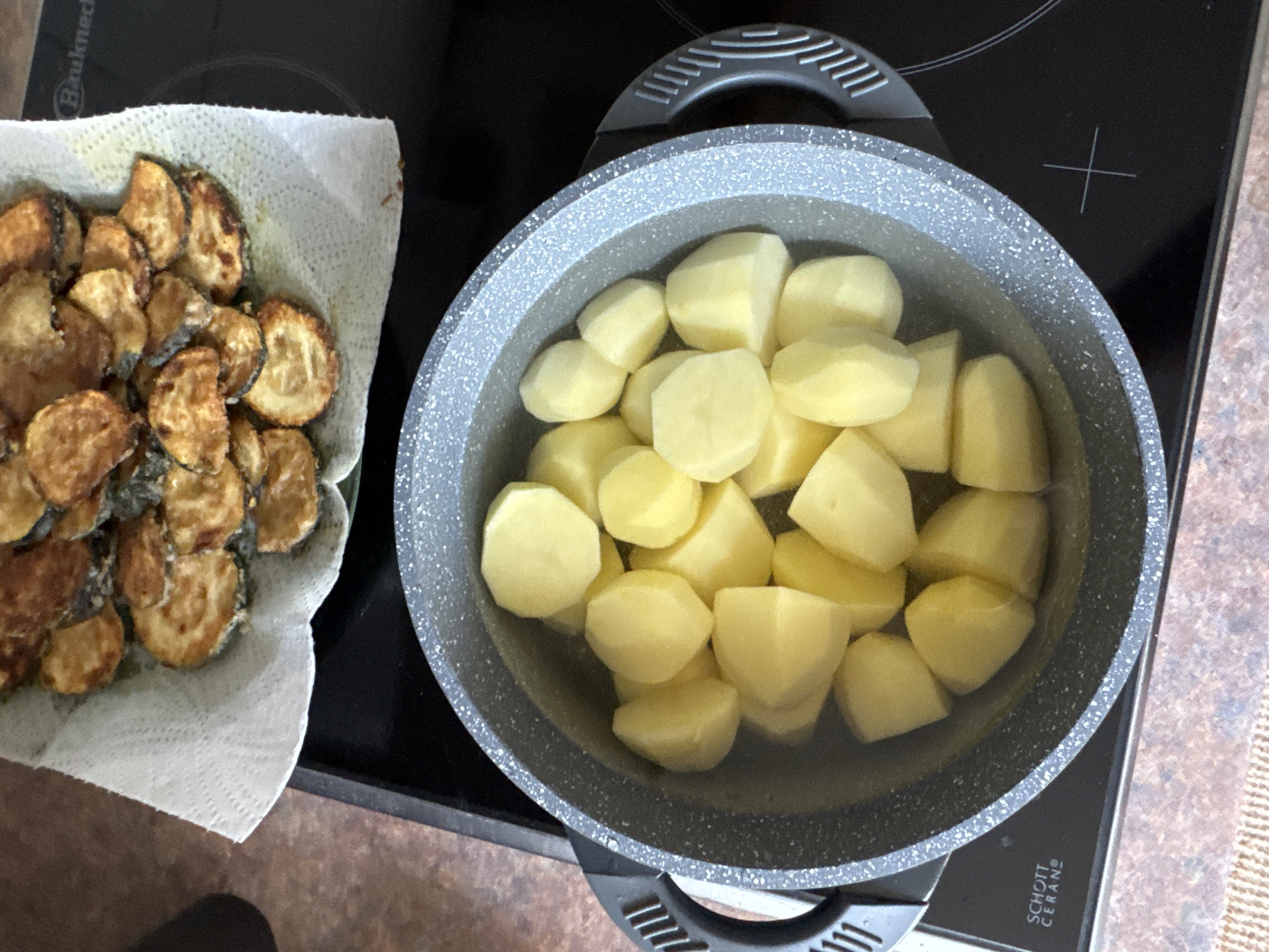 This image depicts a close-up view of food preparation on a black stovetop, likely in a kitchen within Höhenkirchen-Siegertsbrunn, Germany. In the foreground, a grey speckled pot filled with peeled and chopped potatoes submerged in water sits on the stovetop. The pot has black handles. Above and slightly behind the pot, a plate is covered with a white paper towel, and on top of the paper towel are numerous golden-brown, fried slices of what appears to be zucchini or eggplant. The stovetop surface is visible around the pot, with a visible "SCHOTT CERAN" logo indicating the material of the hob, and a heating element symbol. The background is out of focus, showing a brown speckled countertop and part of a dark grey appliance. The scene is well-lit, suggesting indoor lighting.