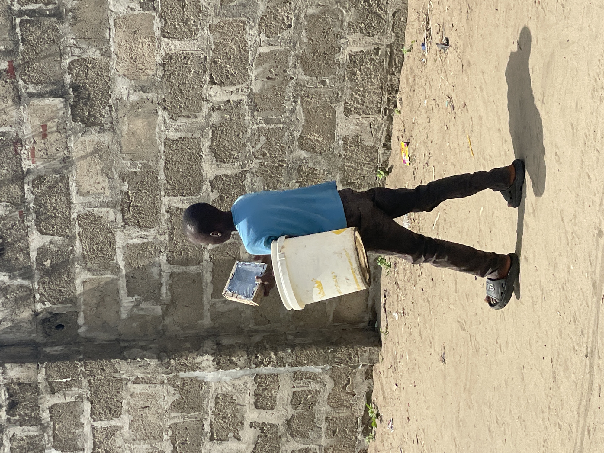 An outdoor, full-body shot depicts a young male walking on a sandy or dirt path in Badagry, Nigeria. He is seen from a side profile, wearing a light blue short-sleeved shirt and dark trousers. On his feet are black slides, one of which clearly displays a white "B" logo on its strap.
In his right hand, he carries a white plastic bucket, which has some yellow staining or discoloration around its rim. In his left hand, he holds a small, rectangular object that resembles a tray or board, covered with a dark substance, possibly paint or cement mix.
Behind him is a textured wall constructed from rough, light-colored blocks or bricks with visible mortar lines. The ground is sandy or dirt-covered, with a few small green plants and a yellow and orange wrapper visible as litter. A long, distinct shadow of the person is cast on the ground to his right, indicating bright sunlight, likely during the early morning or late afternoon. The scene captures an everyday activity, possibly related to construction or a similar manual task.