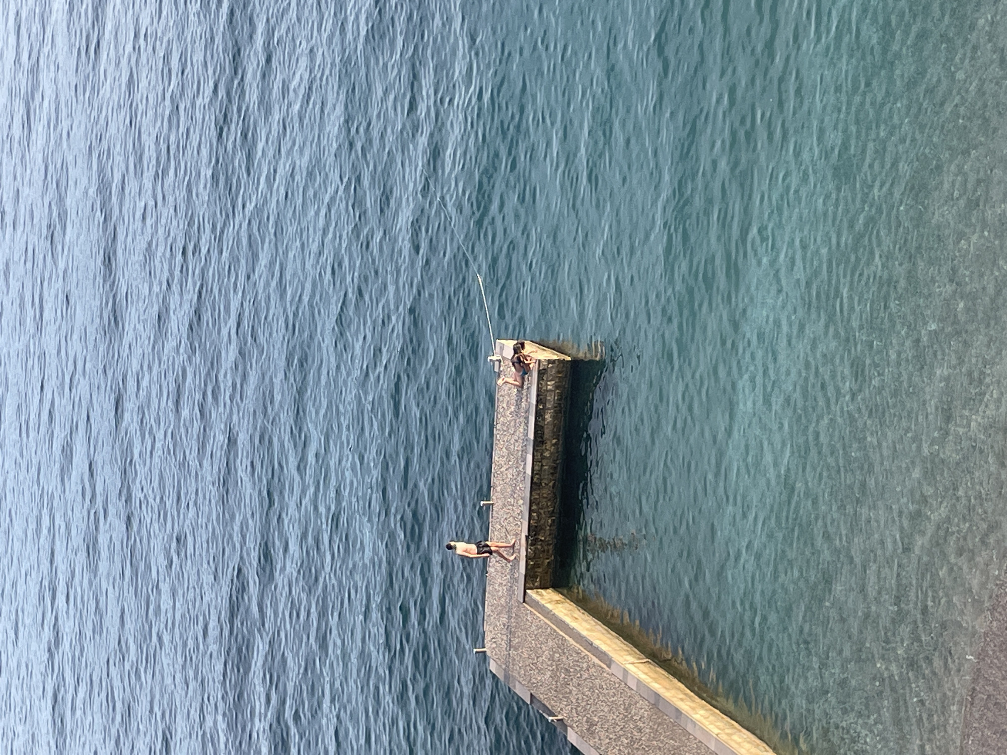 In Budva, Montenegro, a paved stone pier juts into the clear blue sea. Two individuals are visible on the pier. One person stands near the shore end of the pier, facing away from the camera. Another person lies prone on the far end of the pier, their body facing towards the water. The water is a vibrant turquoise, with visible ripples and a sandy bottom beneath the shallow areas. The pier appears to be part of a man-made swimming area or a coastal access point.