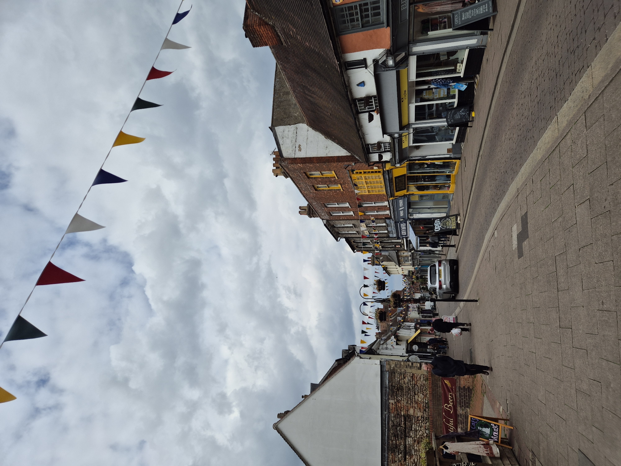 The image depicts a street scene in West Suffolk, United Kingdom. The view is along a narrow street with brick pavement, lined with buildings of varying heights and architectural styles. Many of the buildings appear to be shops or businesses, with storefronts displaying signs and awnings. Overhead, strings of colorful triangular pennants are strung across the street, creating a festive atmosphere. A few people are walking along the street, some appear to be window shopping. A silver car is parked on the road. The sky is overcast with cloud cover.