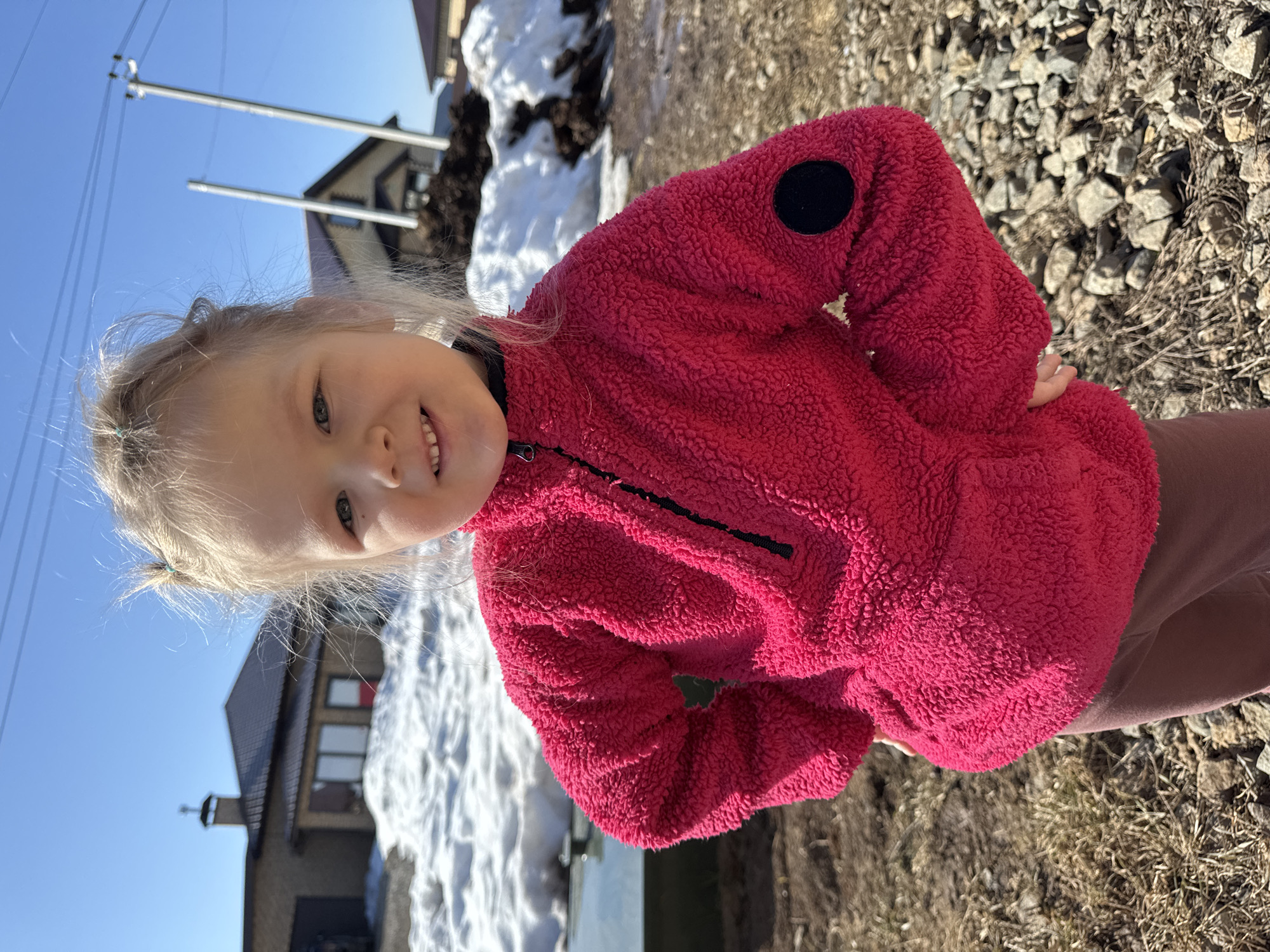 A young child, fair-skinned with light hair tied into two small pigtails, stands centered in the frame, looking directly at the camera with a slight smile. The child is wearing a bright pink, textured fleece quarter-zip pullover with a black zipper and a black circular patch on the upper left sleeve, over dark brown or maroon pants. Both hands are placed on the hips. The foreground consists of bare brown earth, dry grass, and scattered grey rocks. In the midground, several patches of melting snow are visible. The background features residential buildings under a clear blue sky. To the left, a light-colored brick house with a dark sloped roof, chimney (with a cross finial), windows, and a garage is present. To the right, another similar house is partially visible behind mounds of dark soil/debris and snow, along with two tall utility poles carrying power lines. The scene is outdoors in a residential area of Gornyi (Dvurechenskoe S/P), Russia, under clear, sunny conditions.