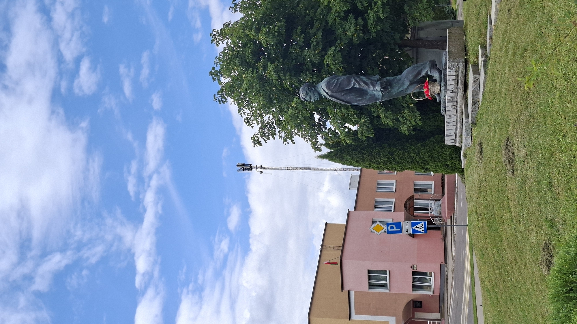 In the city of Myadzyel, Belarus, a bronze statue of a man in a suit stands on a stone pedestal inscribed with "MAKSIM TANK". A basket of red flowers rests at the statue's feet. Behind the statue, a large green tree provides shade, and further back, a multi-story building with pink and beige facades is visible. A cell tower stands tall against a blue sky dotted with white clouds. Traffic signs, including a parking sign and a pedestrian crossing sign, are present near the building. The ground is covered in green grass.