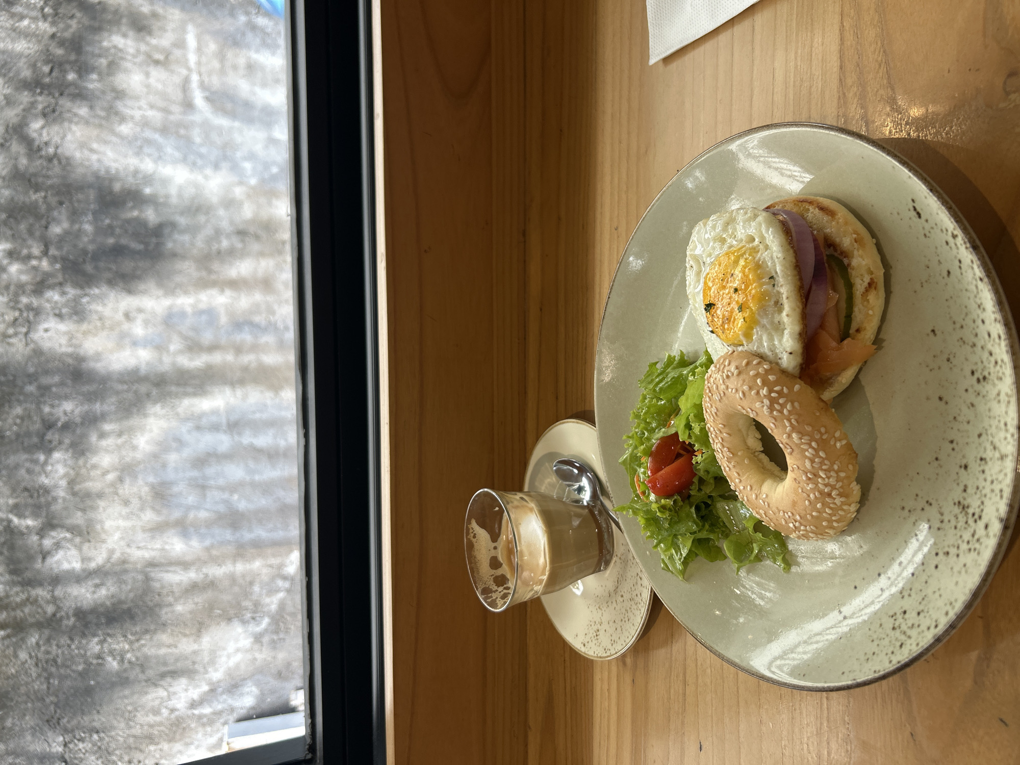 A top-down, eye-level shot shows a plate with a bagel sandwich and a side of salad next to a glass of coffee, set on a wooden table adjacent to a window. The bagel sandwich features a fried egg, red onion, and salmon between the two halves. The salad is composed of lettuce and tomato. The coffee glass is partially filled with a light brown liquid and sits on a matching saucer. Outside the window, there is a blurry, gray scene, potentially indicating a hazy or overcast day. The location is identified as Kuta Selatan, Indonesia. No people are visible in the image.