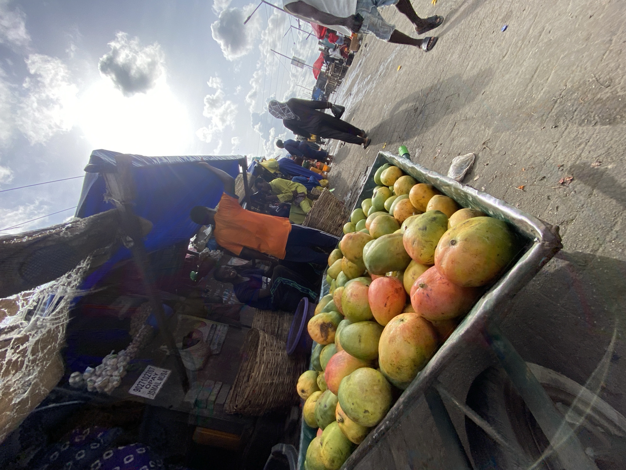 The image depicts a bustling street scene, likely a market, in Lagos, Nigeria. In the foreground, a cart overflows with mangoes, varying in ripeness from green to red-orange. People walk along the side of the road adjacent to stalls covered with blue and yellow tarps. Near the cart in the foreground are several people, some standing and others walking. In the distance, rows of shops or stalls are visible. The sky is bright with scattered clouds. A white sticker, likely a vendor’s tag, reads "9027102707 OPAY ISMAIL MUSA".
