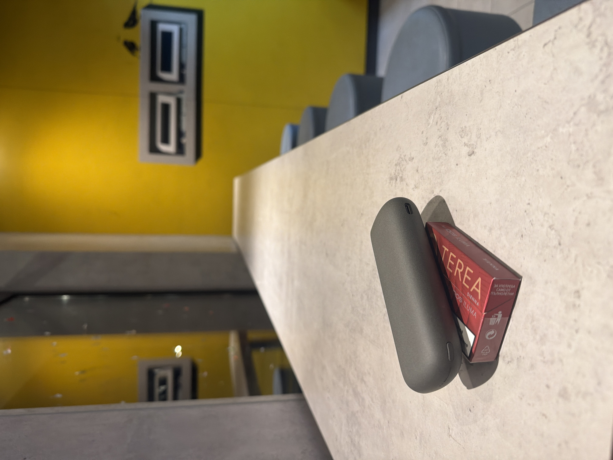 This media file, originally rotated 90 degrees clockwise, depicts a modern indoor scene, likely in Sofia, Bulgaria, based on provided context and visible text. The perspective is slightly elevated, looking down over a light-colored, textured counter or ledge that diagonally crosses the frame from lower left to upper right.
In the foreground, resting on this counter, is a dark grey, smooth, oblong electronic device, possibly a heated tobacco device. Next to it stands a small, dark red rectangular box. The box prominently displays "TEREA" and "SIENNA," with "FOR ILUMA" visible below. On its side, Cyrillic text reads "ЗА УПОТРЕБА САМО ОТ ПЪЛНОЛЕТНИ" (FOR USE ONLY BY ADULTS), accompanied by a "do not litter" symbol and a recycling symbol, indicating it's a tobacco product.
Behind the counter, in the midground, several grey, rounded, upholstered seats or stools are arranged in a row, suggesting a seating area. The background features a striking, solid yellow wall. Mounted on this wall are two stacked, dark-framed rectangular elements with light, reflective interiors, resembling modern light fixtures or decorative panels. Below the counter, a reflective surface, possibly a polished floor, shows a distorted reflection of the yellow wall and the lower rectangular wall fixture.
The overall impression is a clean, contemporary setting with minimalist design elements, characterized by the stark yellow wall, grey seating, and textured counter. The lighting appears to be artificial, typical of an indoor environment. No people are visible, and there are no specific activities taking place, creating a sense of quiet observation.