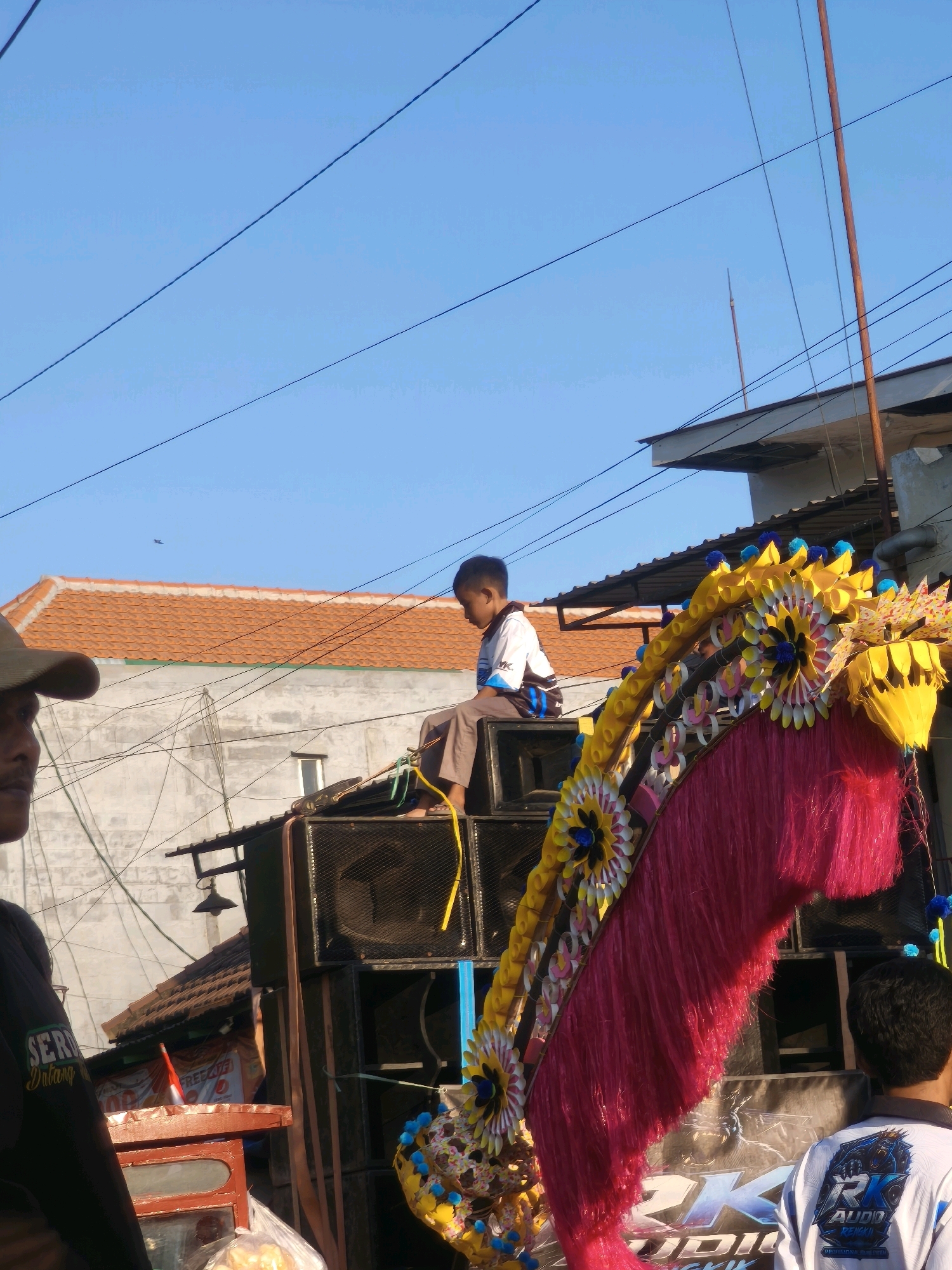 A young boy is seated on top of large black speakers, looking to his left. He is wearing a white shirt with blue and black accents and brown pants. To his left, a man wearing a tan baseball cap and a black t-shirt with green lettering is partially visible, looking to his right. In the foreground, a brightly colored, elaborate decoration with pink fringe and yellow and blue floral elements is prominent. Behind the speakers, a building with a red tiled roof and a light-colored wall is visible. The scene appears to be outdoors, with a clear blue sky above and power lines crisscrossing the frame. The image was taken in Kepuh Kiriman, Indonesia.