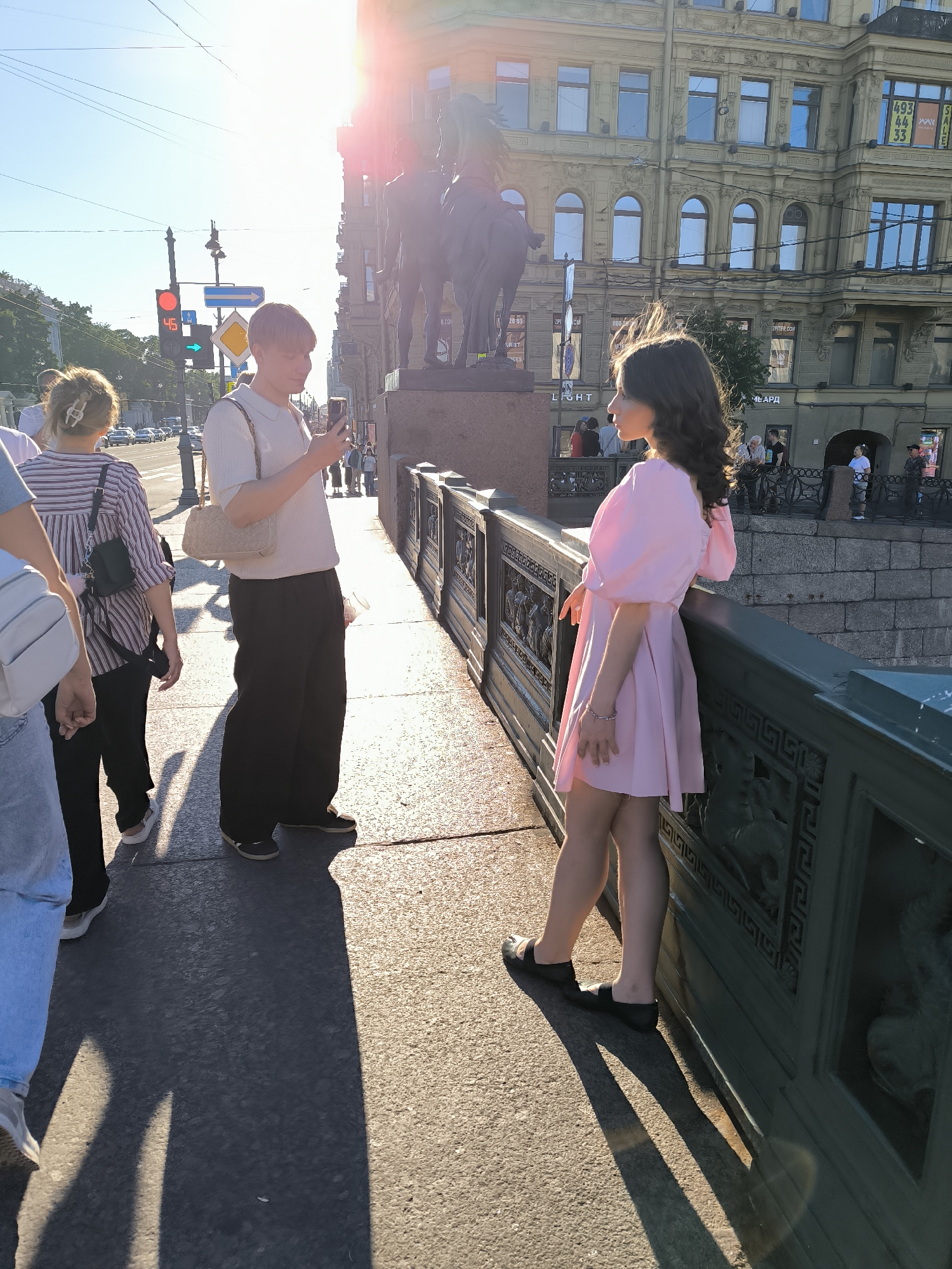 This image captures a sunlit scene on the Anichkov Bridge in Saint Petersburg, Russia, likely during late afternoon due to the strong, low-angle sunlight and long shadows.
In the foreground right, a young woman with dark, wavy hair, wearing a light pink, short-sleeved dress with puffed shoulders and black flat shoes, stands leaning against the bridge's dark green, ornately detailed metal railing. She faces left, with her left hand resting on the railing, revealing a silver bracelet on her wrist. To her left and slightly behind her, a young man with light brown hair, dressed in a light-colored polo shirt, dark wide-leg trousers, and carrying a light beige shoulder bag, stands facing forward while looking down at a smartphone held in his hands.
Further left, partially visible from the back, is another person with light brown hair secured by a claw clip, wearing a horizontally striped shirt and dark trousers, and carrying a black shoulder bag. Their lower left leg is obscured by a lighter-colored pant leg, possibly belonging to another person.
Behind the young man, the base and part of a large equestrian statue, consistent with the "Horse Tamers" sculptures of the Anichkov Bridge, are visible. The bridge's pedestrian path, a light-colored pavement, shows prominent long shadows cast by the subjects and the railing, extending towards the left.
In the middle ground, a red traffic light displays the number "45" (a countdown timer), positioned next to a blue arrow street sign pointing right and a yellow diamond-shaped sign. Beyond the bridge, classical-style buildings with numerous windows line the street. One building in the background right features faint signage, including "SALE" and a partial phone number "493 44 33," typical of local establishments. The sky is bright and clear, with a significant lens flare from the sun positioned in the upper right quadrant of the image.