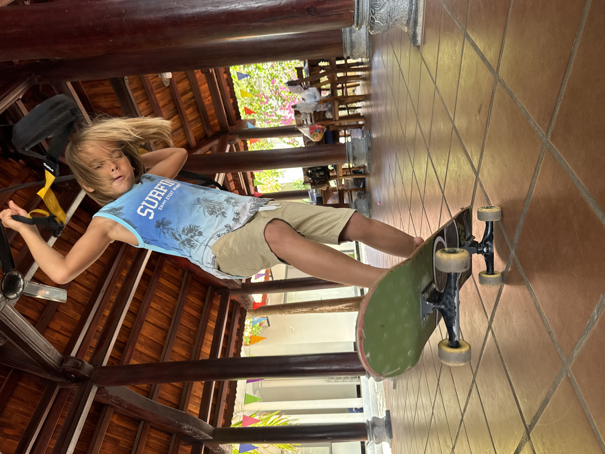 A young boy with blond hair is lying on a green skateboard in what appears to be a public space in Mũi Né, Vietnam. The boy is wearing a blue tank top with the words "SURFING ENJOY EVERY MOMENT" printed on it, and khaki shorts. He has his arms raised behind his head, and his face is relaxed. The skateboard is resting on a tiled floor, and the background shows a covered walkway with wooden pillars and beams. Through the pillars, there is a view of a garden with colorful decorations, including pennants. There are chairs and tables in the background, suggesting a cafe or restaurant.