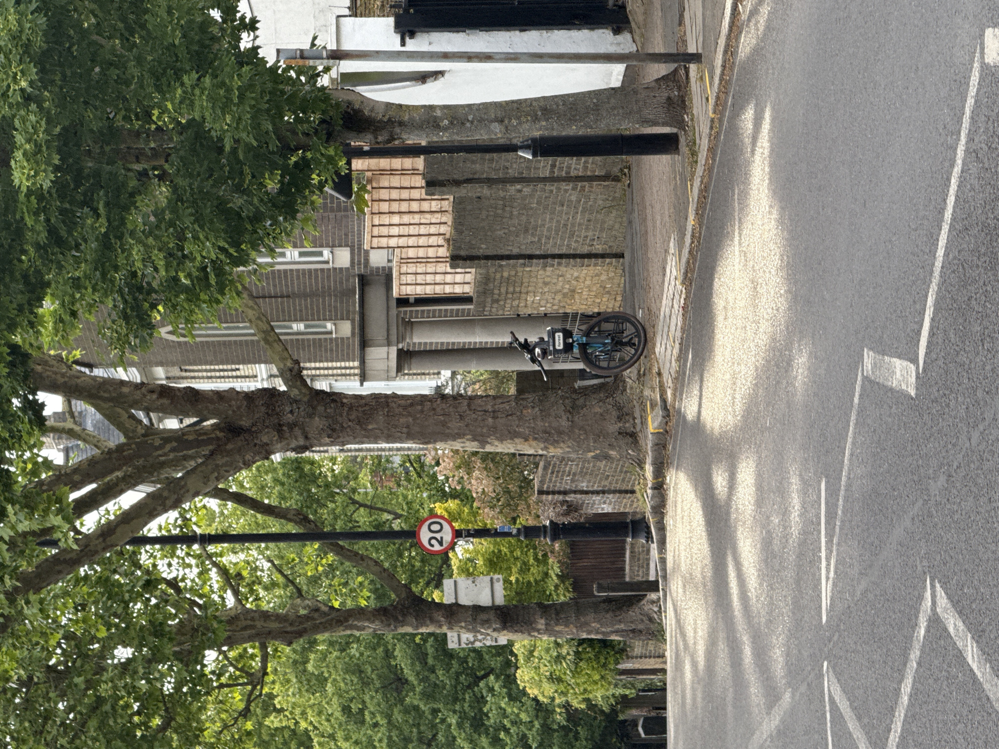 The image depicts a street scene in London, United Kingdom. A bicycle is parked along the side of the road, near a row of houses. A 20 mph speed limit sign is visible on a lamppost, partially obscured by tree branches. The road surface is marked with white lines. Sunlight casts shadows on the road. The scene is typical of a residential street in London, characterized by mature trees and traditional brick buildings. No people are visible in the image.