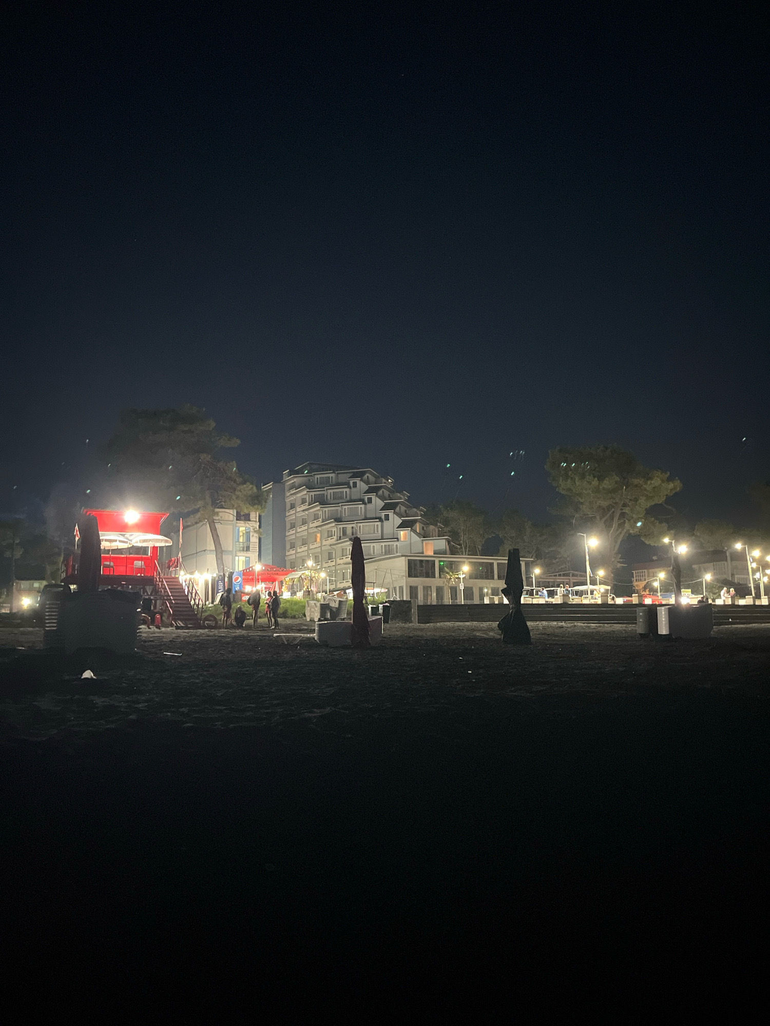 The image depicts a nighttime scene on a beach in Ureki, Georgia. In the foreground, the dark sand of the beach is visible, with some beach furniture such as lounge chairs and umbrellas scattered about. Further back, a red structure with illuminated stairs and a canopy stands out, with a few people gathered around it. Behind this structure, a large, multi-story white building with many windows is illuminated, suggesting it is a hotel or resort. Several trees are present in the midground, and streetlights are visible, illuminating the area and casting light on the building and the beach. The sky above is dark, indicating it is nighttime.