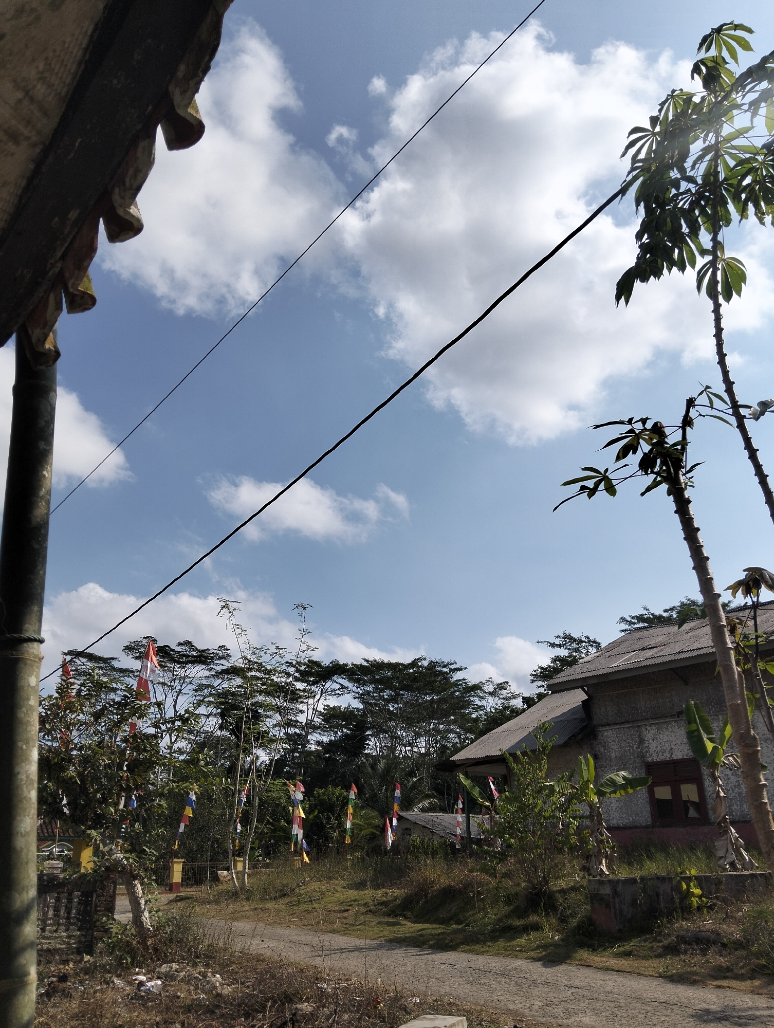The image depicts a rural scene in Karangnunggal, Indonesia, under a bright blue sky with scattered white clouds. A narrow, unpaved road runs through the center of the frame, leading towards a modest, weathered house with a corrugated metal roof. The house appears to have a reddish-brown window frame. In the foreground, to the left, a thick, dark cylindrical pole, possibly made of bamboo, is partially visible, supporting an overhanging roof structure made of aged material.

Along the roadside, a variety of vegetation is present, including trees, bushes, and dry grass. Several tall poles are adorned with colorful triangular flags, alternating between red and white and other hues like blue and yellow, suggesting a festive occasion. Two tall, slender trees with large, palmate leaves, possibly cassava plants, stand on the right side of the frame, reaching towards the sky. A dense line of trees forms a backdrop across the horizon. The overall impression is one of a quiet, rural setting, possibly during a local celebration. No people are visible in the image.