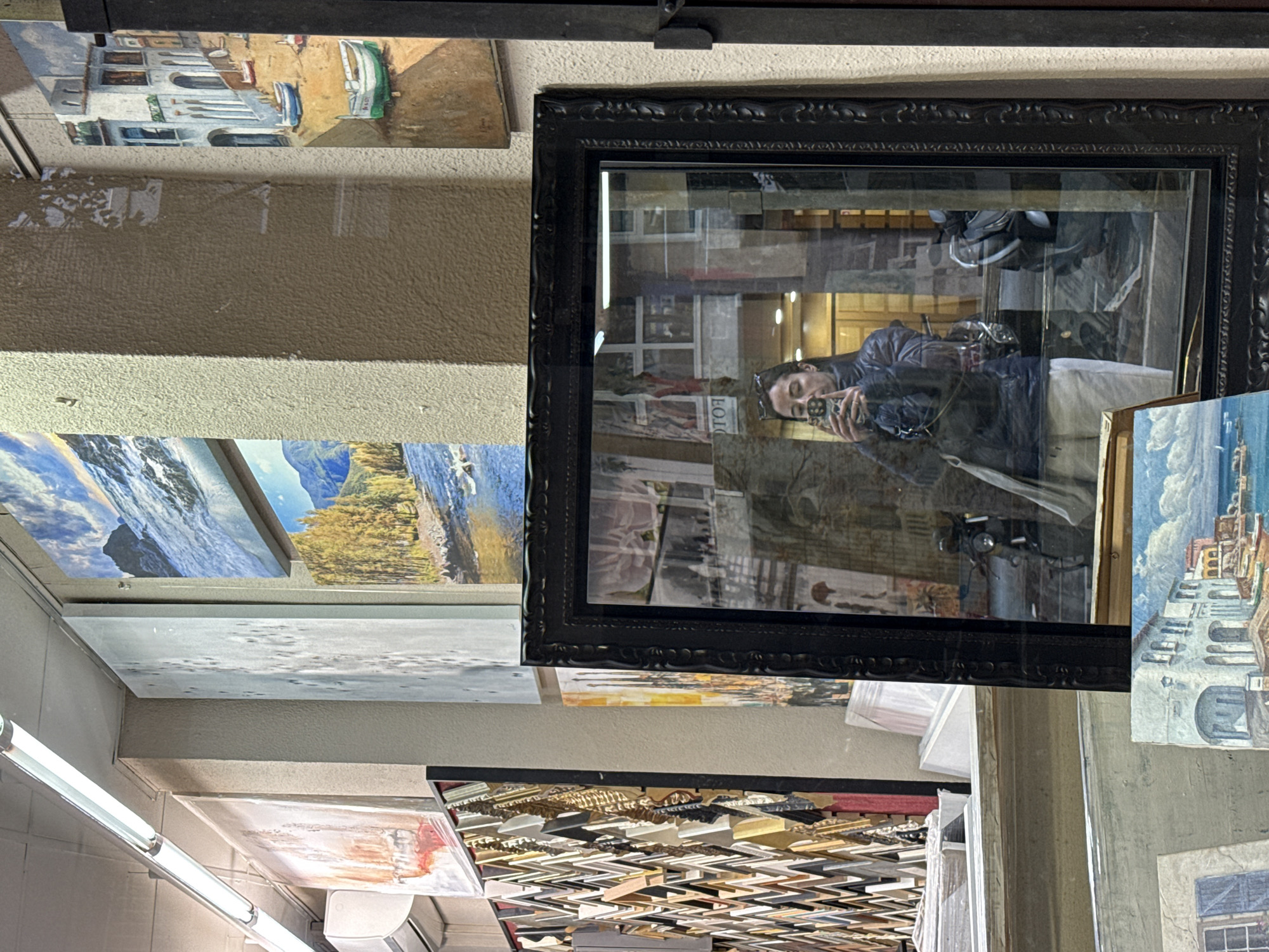 The image is taken indoors, appearing to be an art store or gallery in Barcelona, Spain. Paintings and framed artworks are densely packed, suspended from the ceiling. There are paintings depicting seascapes and coastal towns with whitewashed buildings. One notable element is a large, ornate black-framed mirror reflecting a person holding a smartphone to take a picture, as well as their backpack and other objects in the surrounding environment. Below the hanging art, there's a packed array of wooden picture frame components. Fluorescent lights are visible.